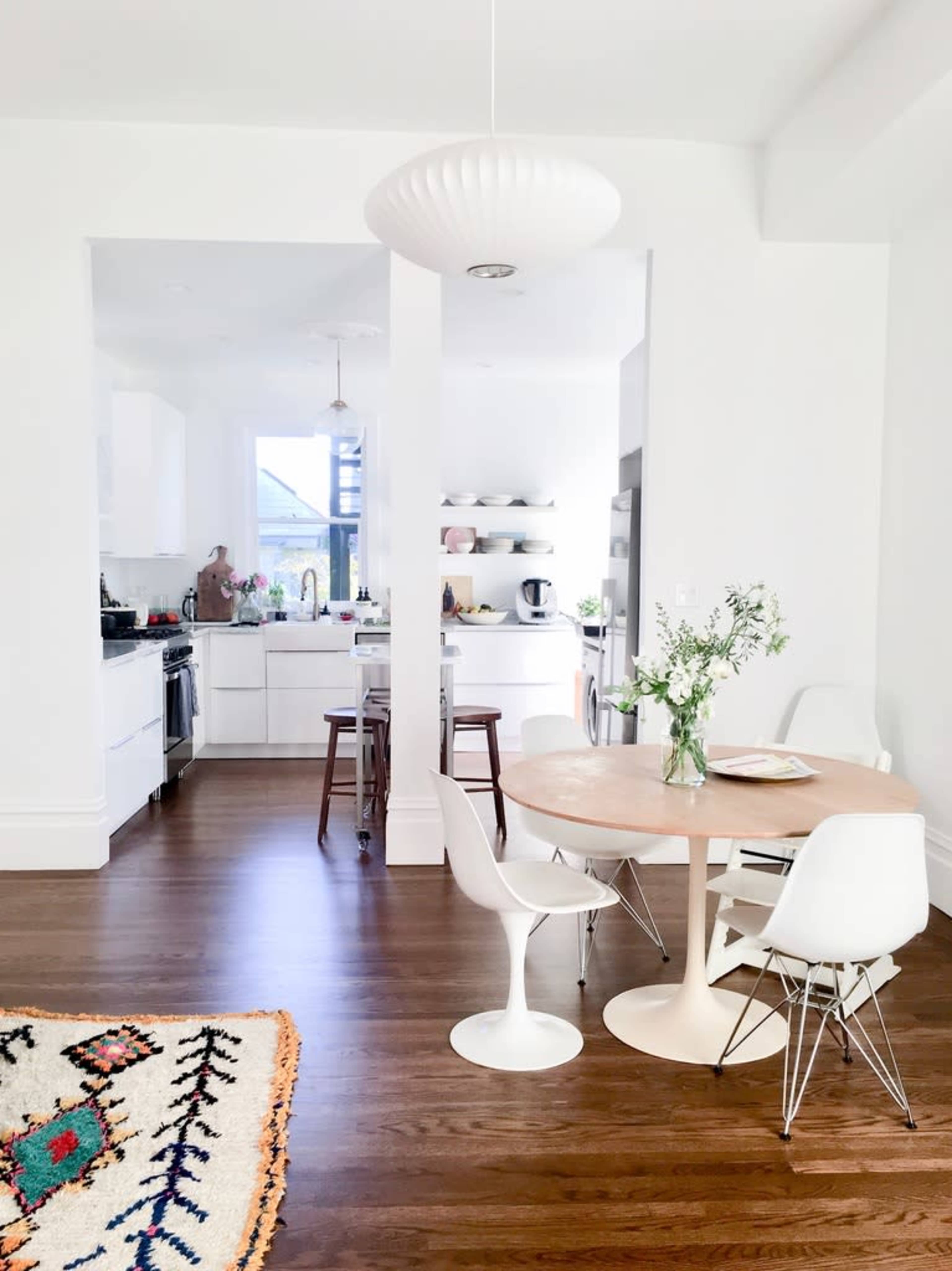 The image depicts a bright and modern kitchen and dining area with a circular table, white chairs, and a view of a well-organized kitchen workspace in the background.