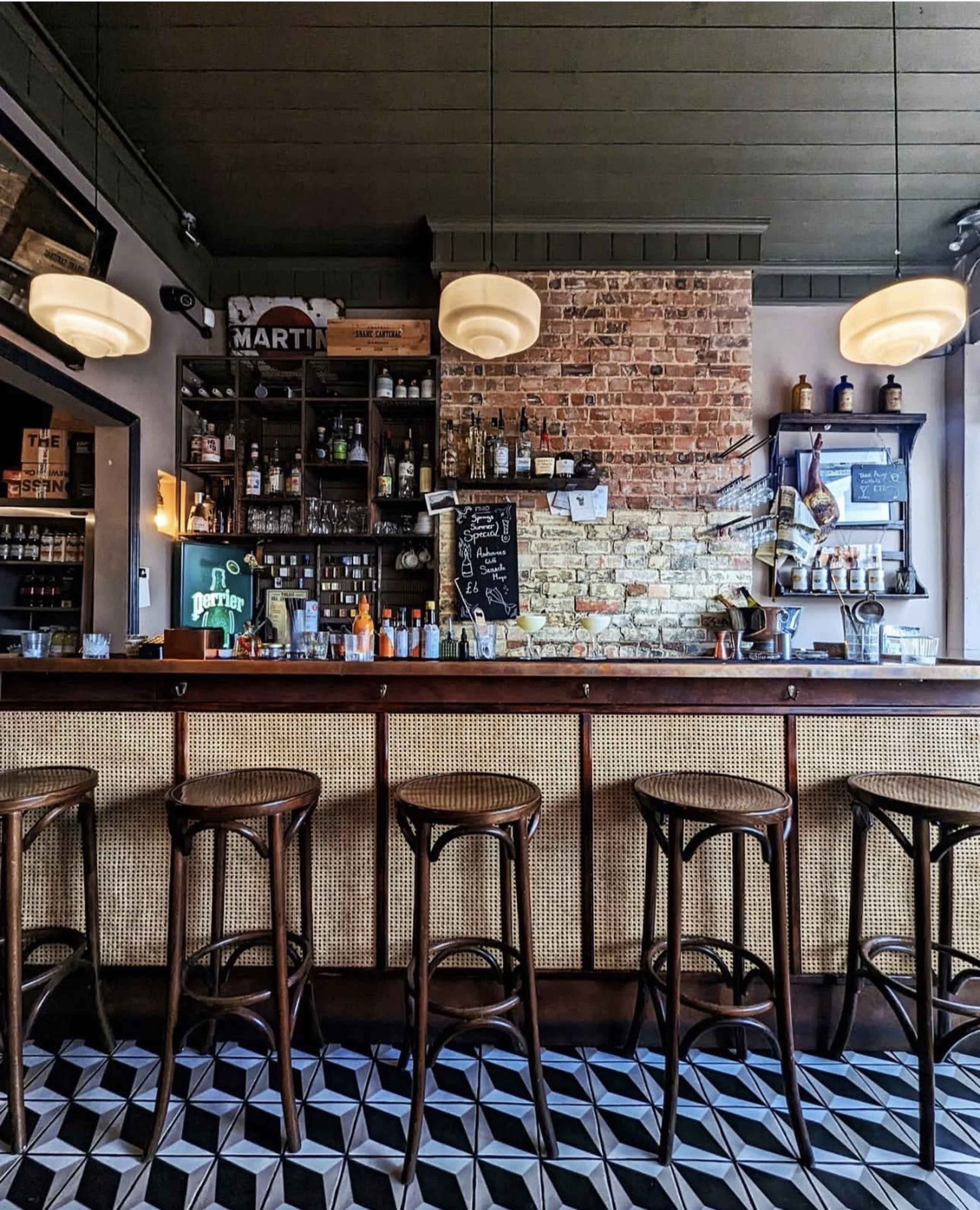 The image shows a bar interior featuring a wooden counter with high stools, a brick wall behind the bar, and various bottles displayed on shelves.