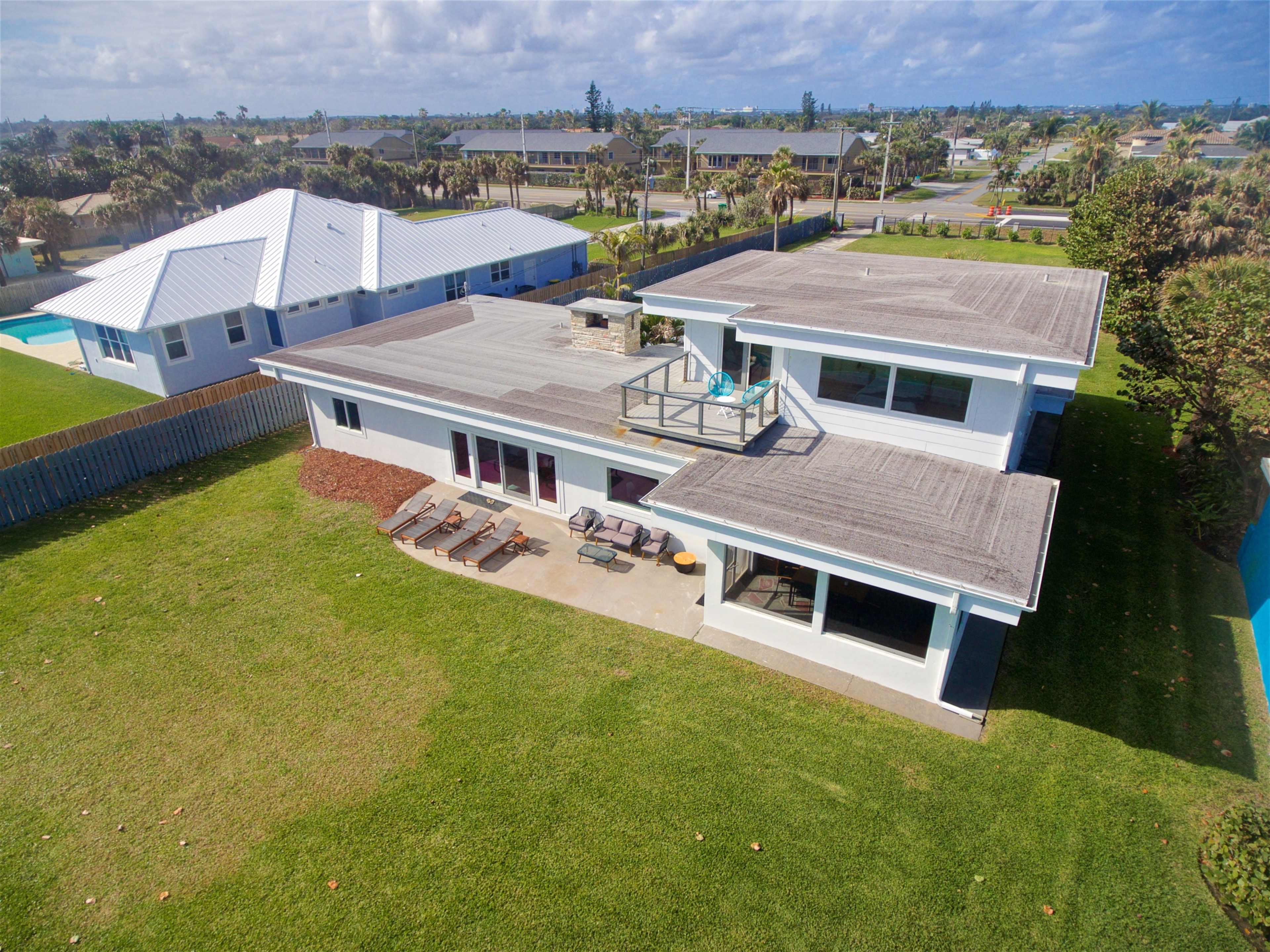 A two-story, modern house with a flat roof and a patio area, surrounded by green grass and neighboring properties in a residential area.