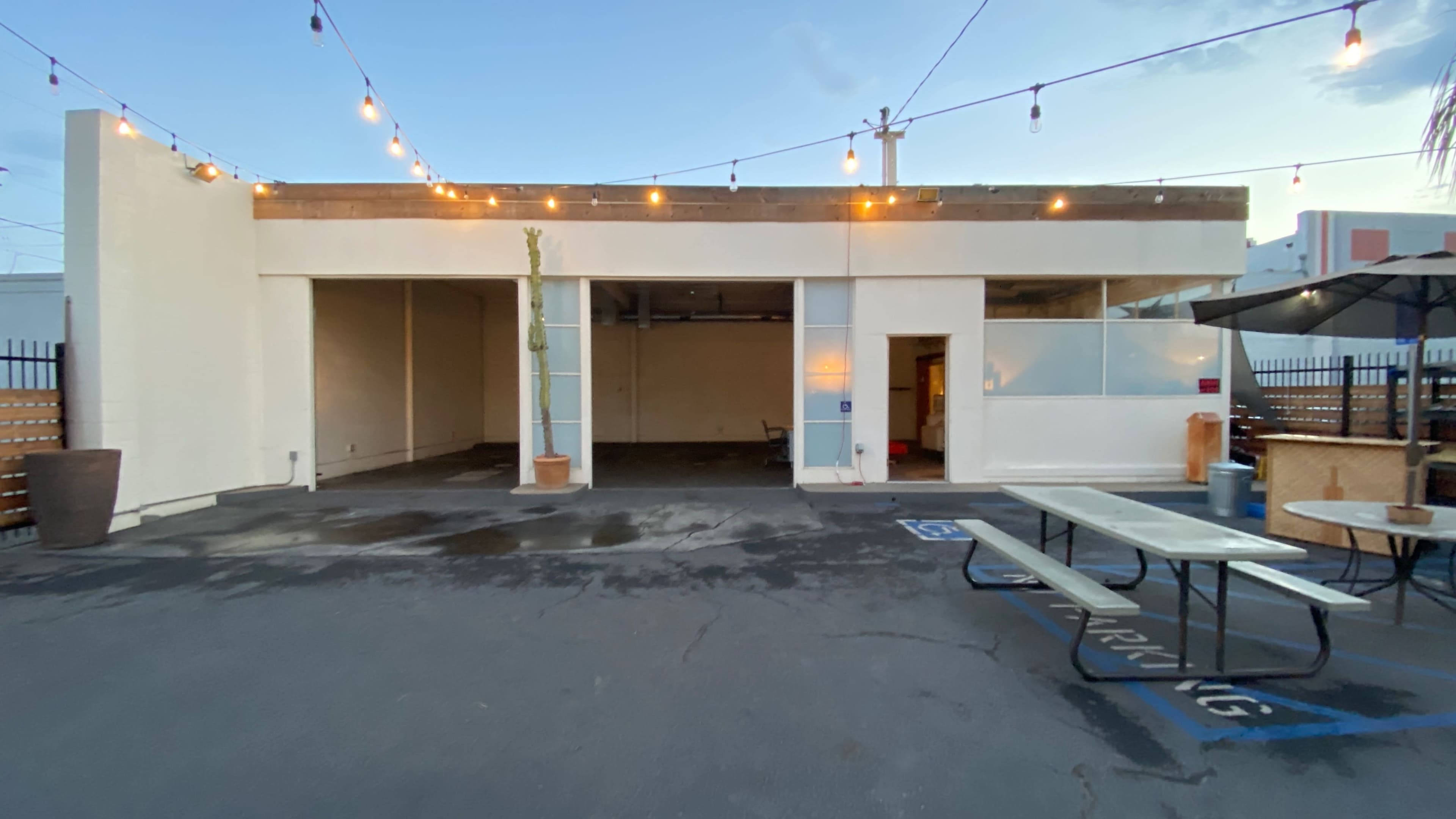 An open outdoor space features a building with string lights overhead, a concrete surface, and a picnic table set against a minimalist backdrop.