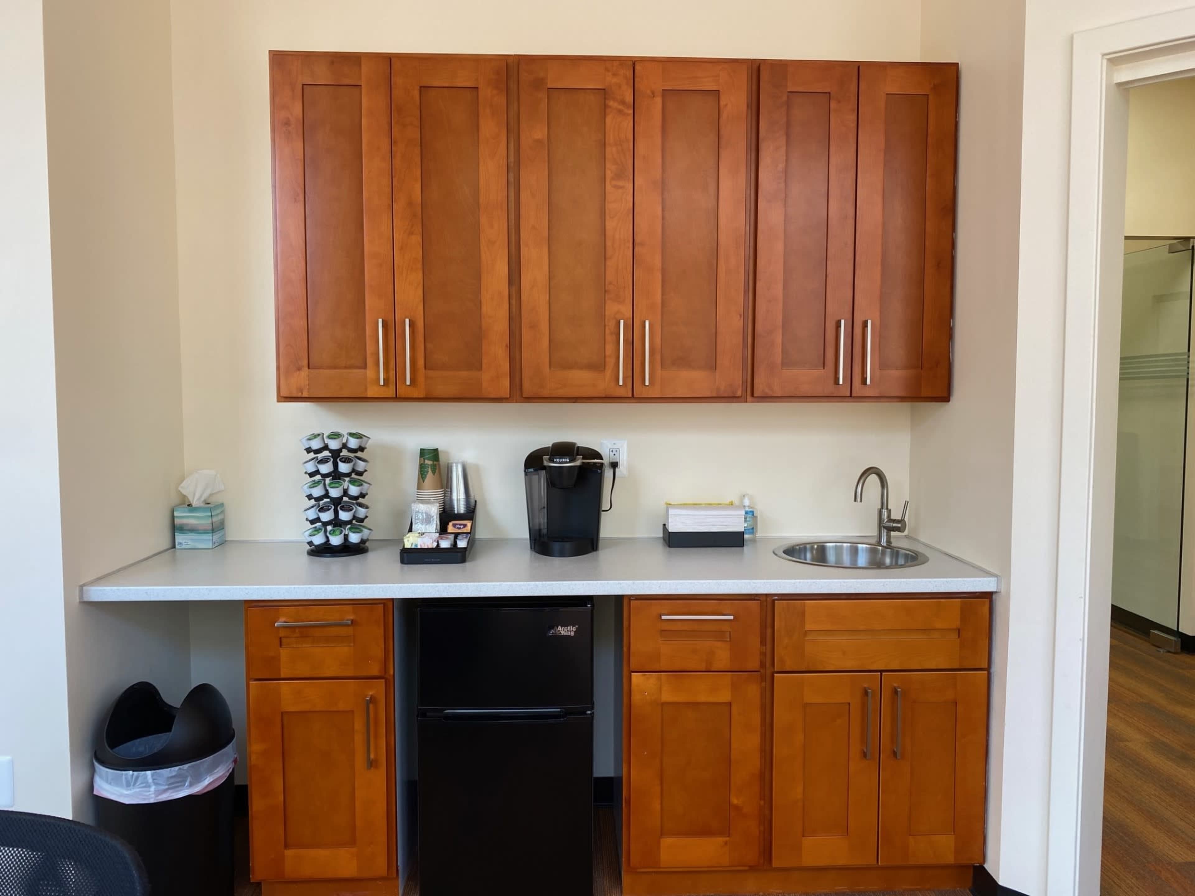 A kitchenette with wooden cabinets, a countertop, a sink, a coffee maker, and a small refrigerator.