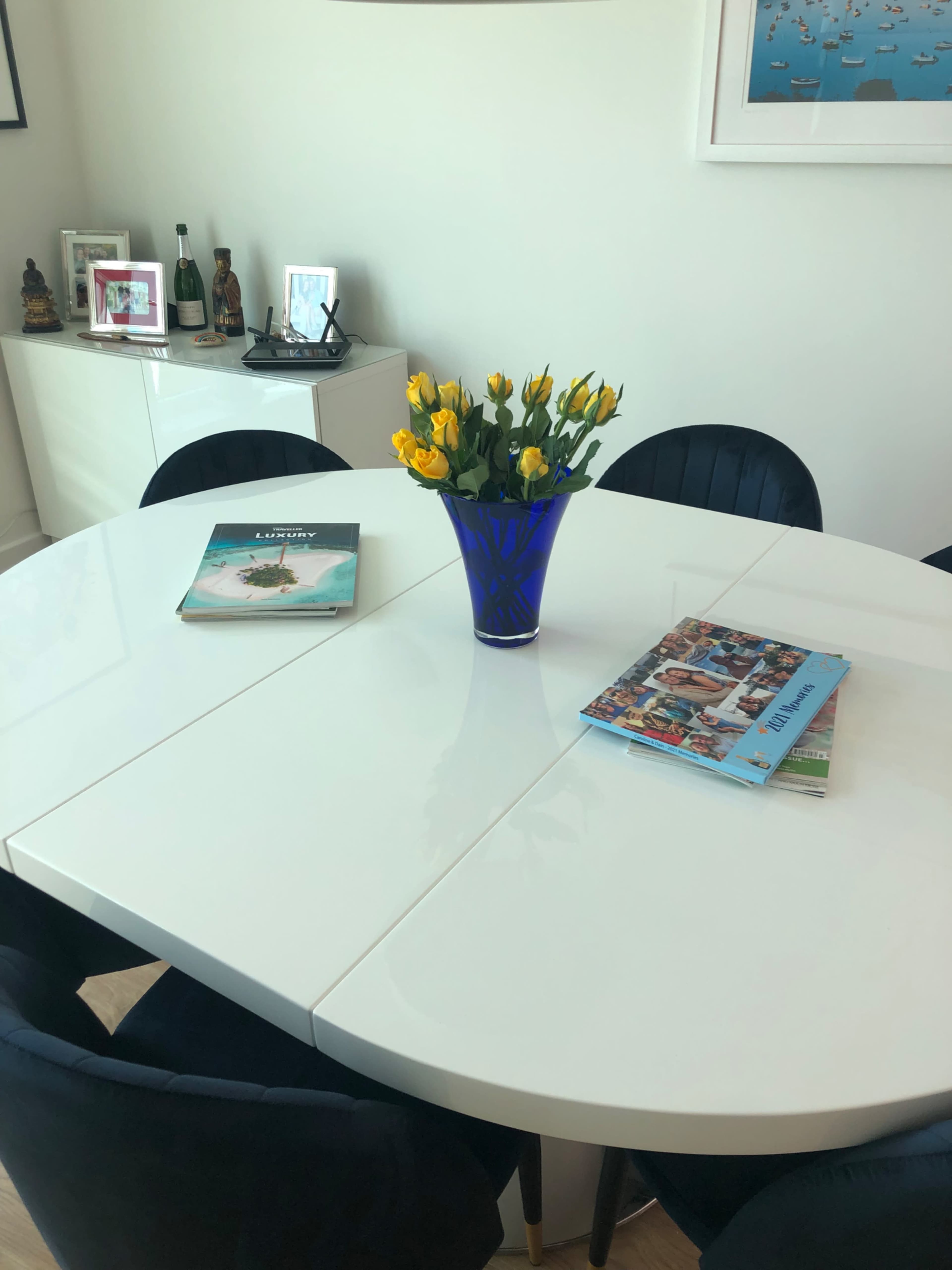 A white dining table is set with two magazines and a blue vase of yellow flowers in the center, surrounded by black chairs.