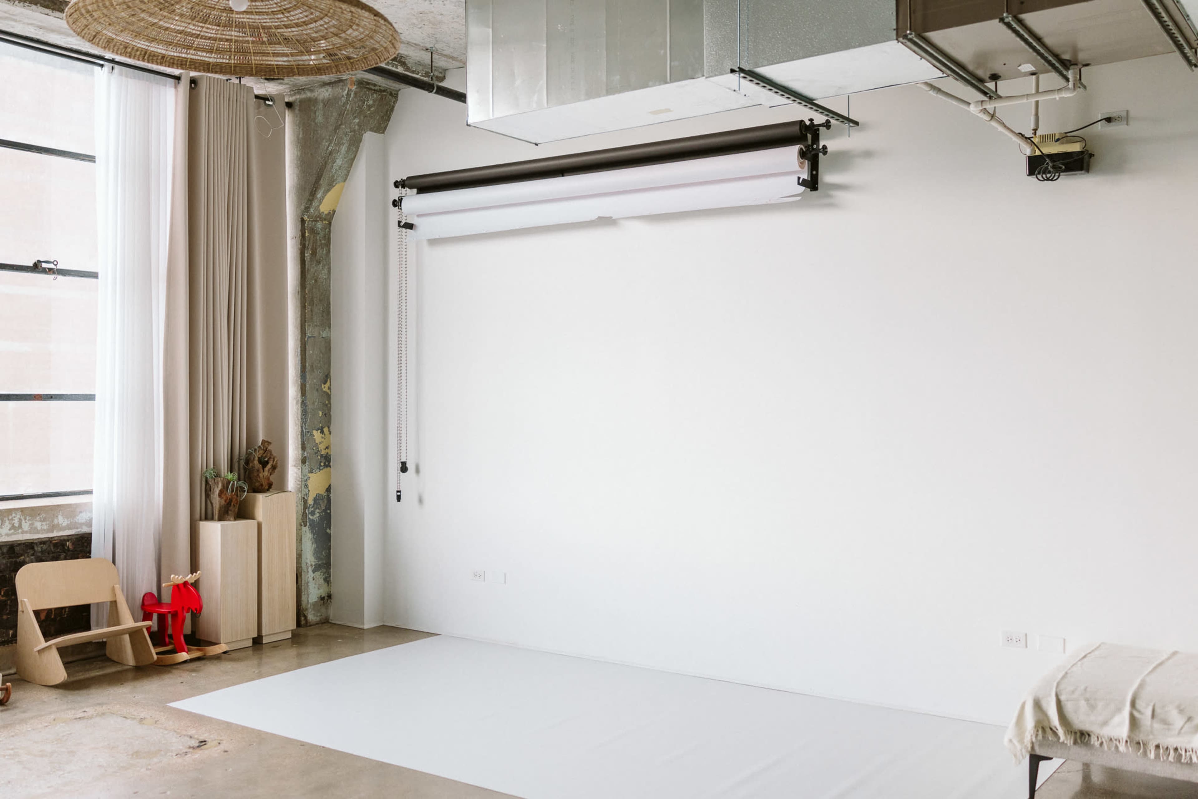 The image shows a minimalist photography studio with a white backdrop, exposed concrete walls, and natural light streaming through a large window.