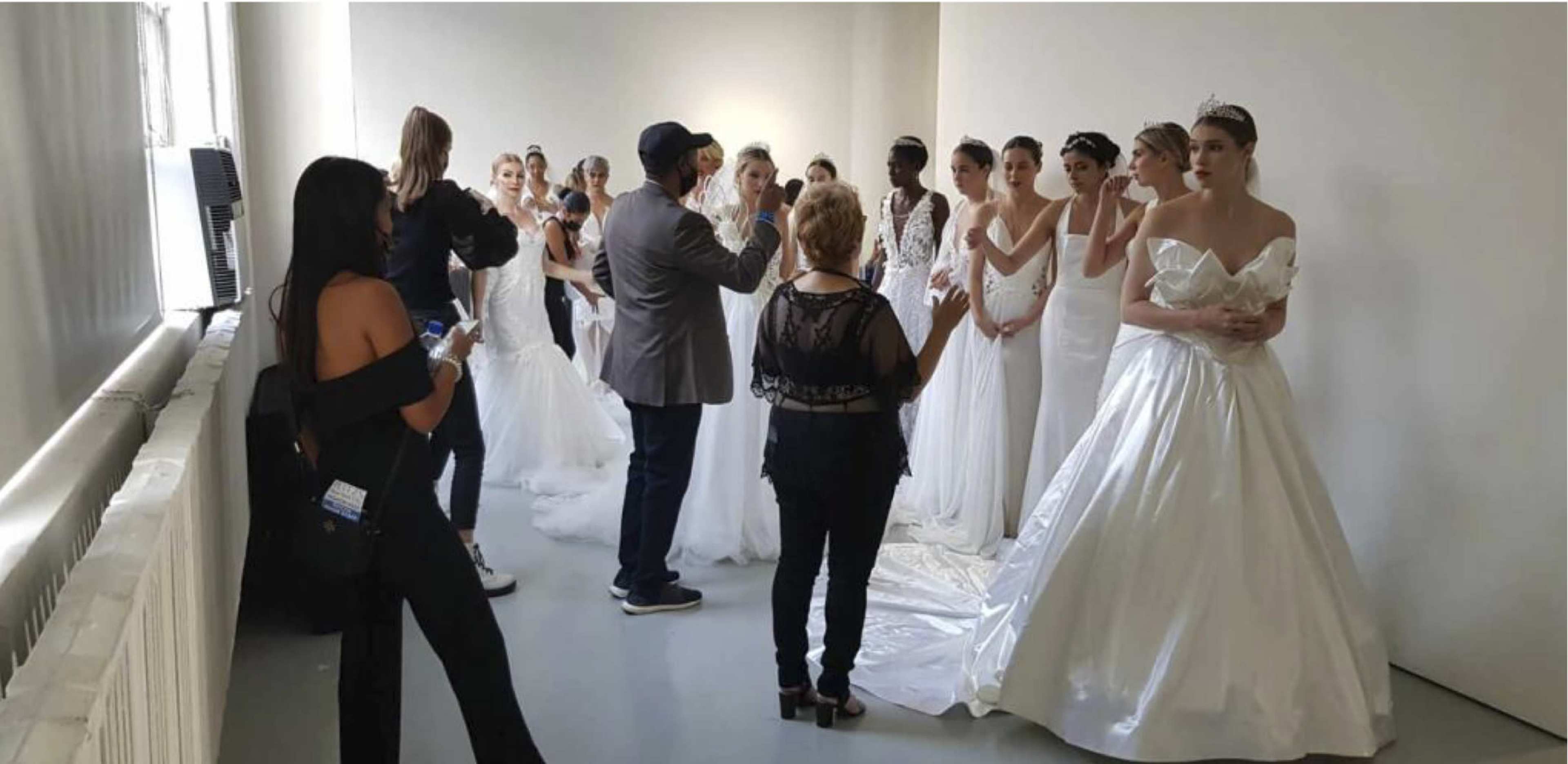 A group of models in wedding gowns stands in a light-filled room while a designer or stylist gives instructions to them.
