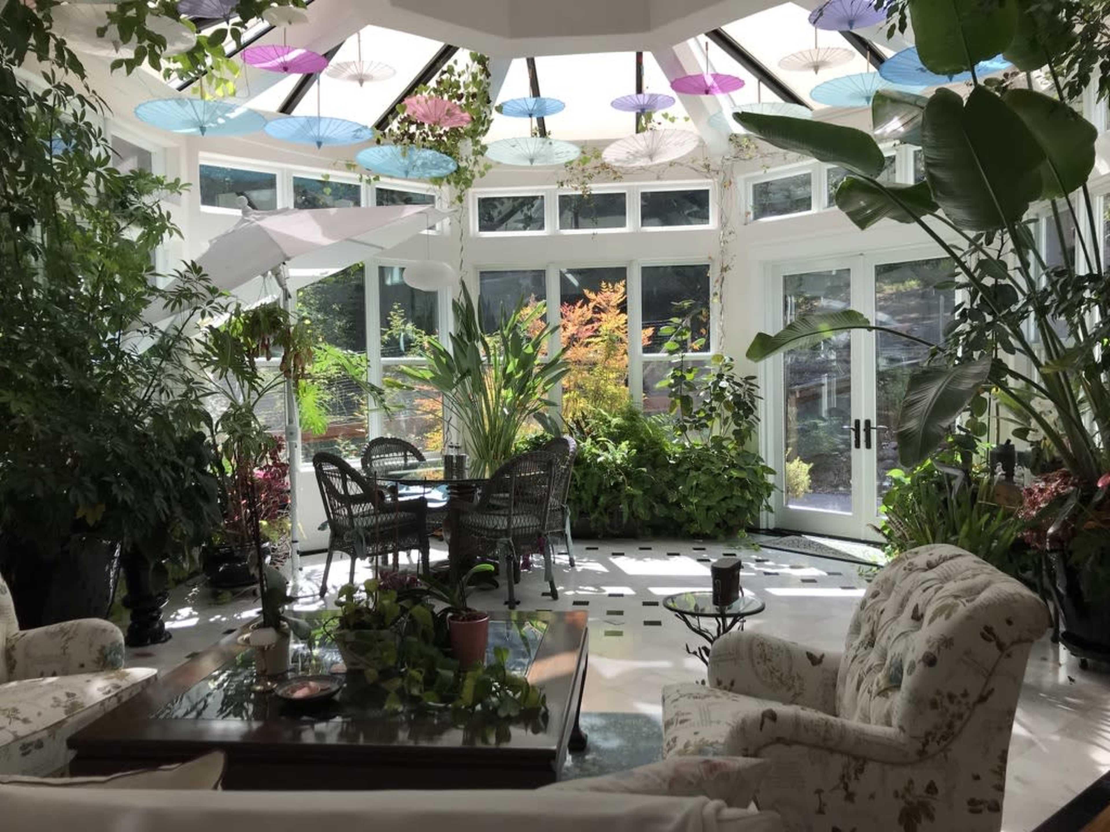 The image shows a bright sunroom filled with various indoor plants, wicker furniture, and colorful hanging decorations.