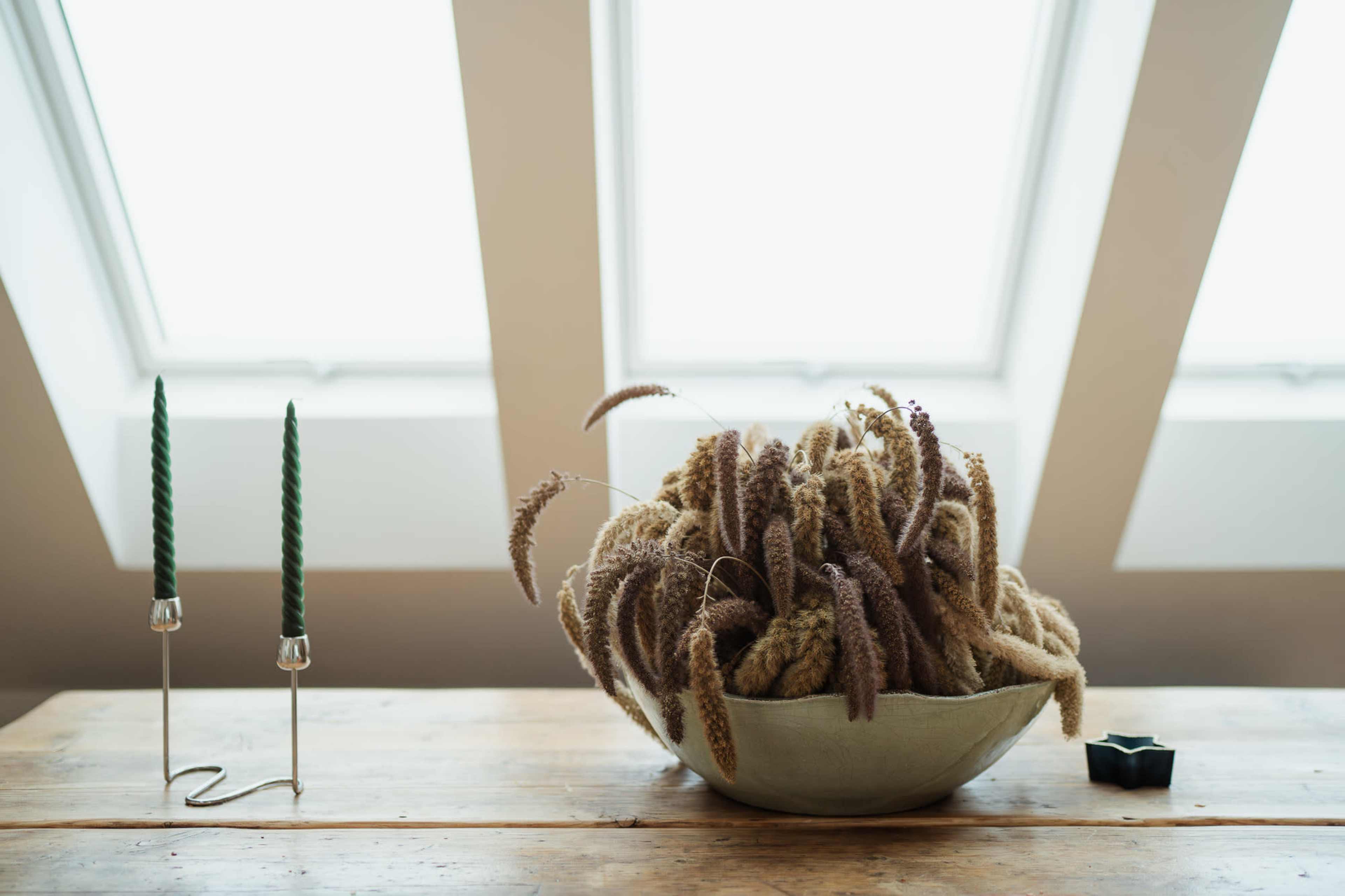 A bowl filled with dried grasses sits on a wooden table alongside two green candlesticks.