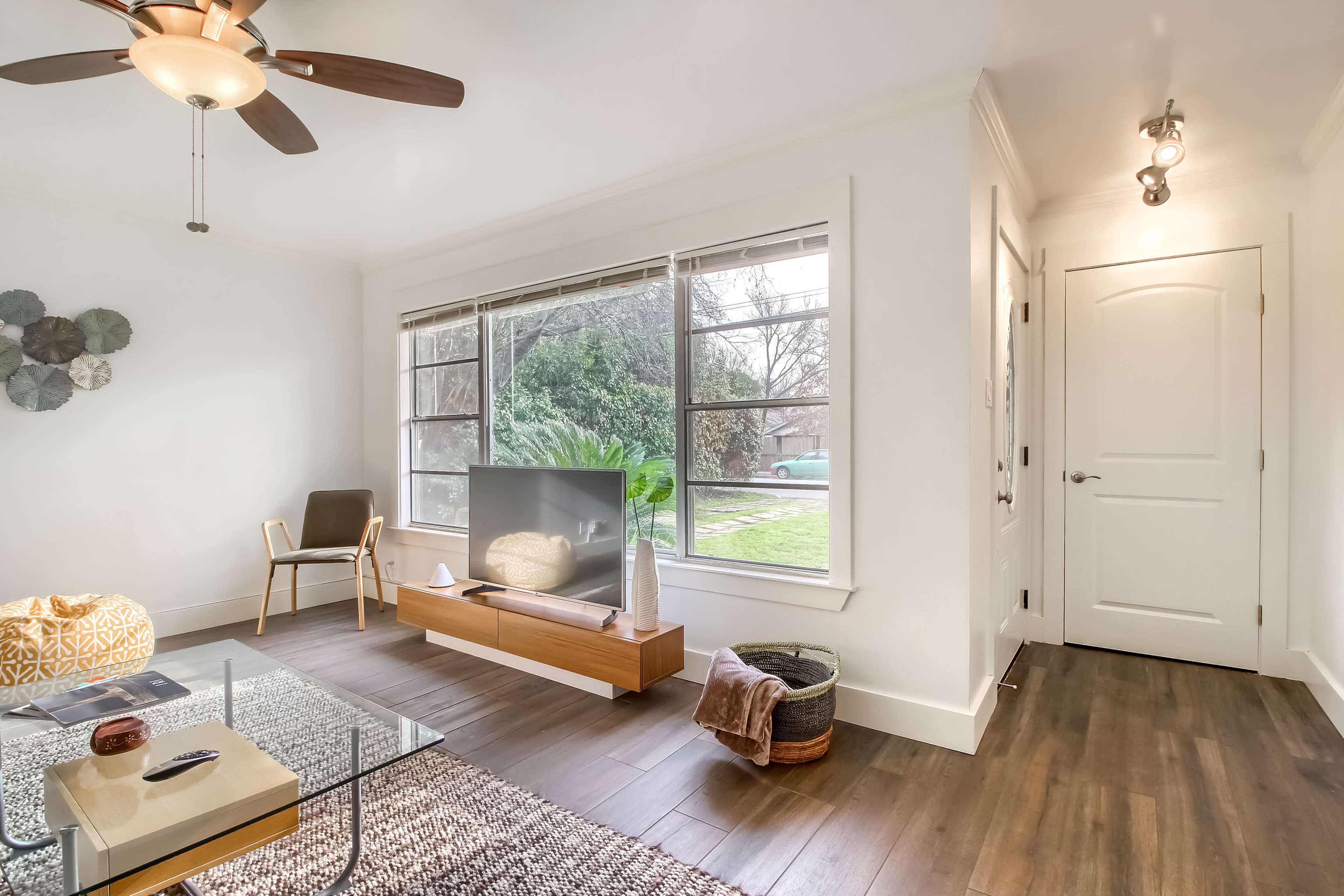 A modern living room with a large window, a television on a low wooden console, a glass coffee table, and a chair in one corner.