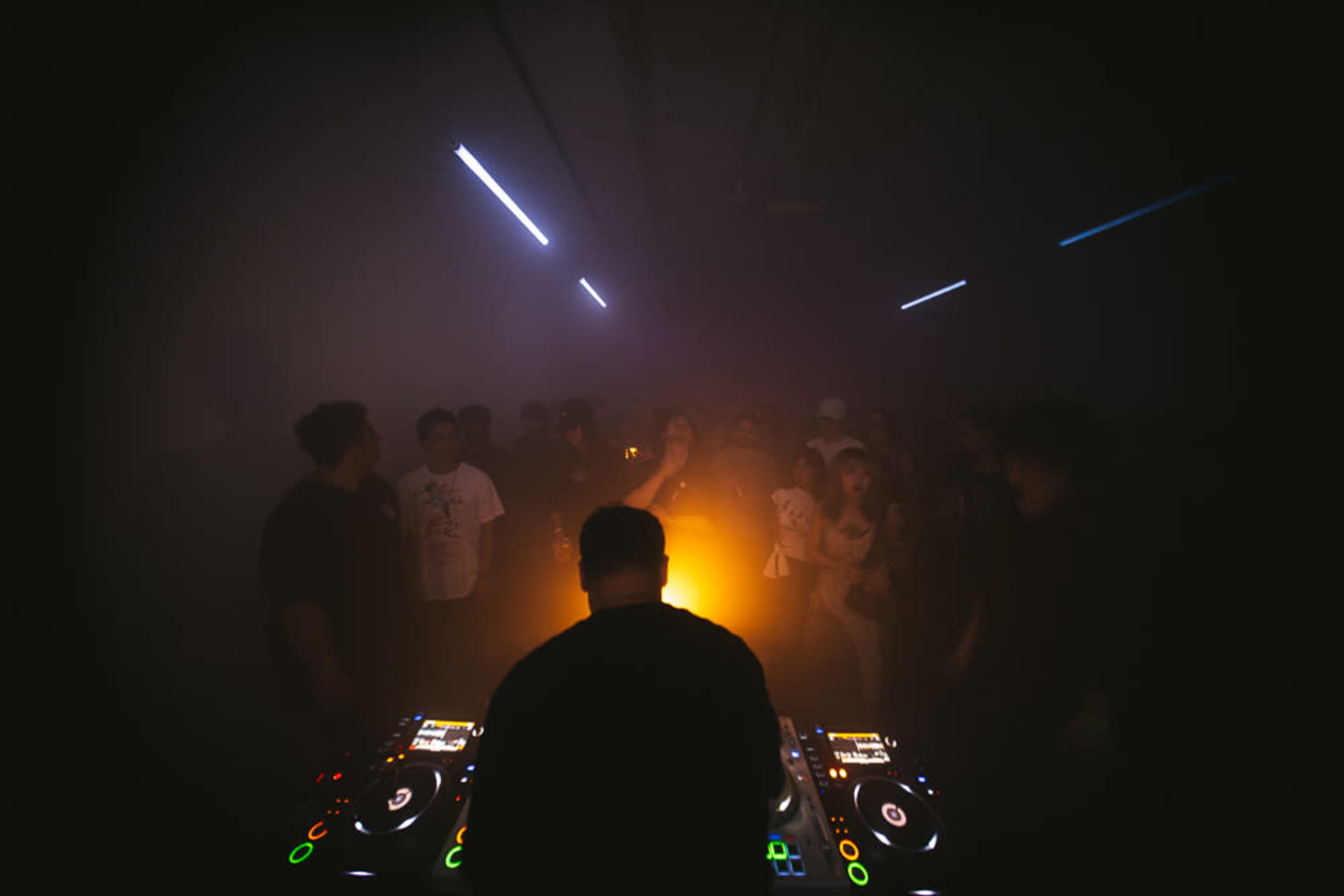 A DJ stands at a control table in a dimly lit room, with a crowd of people dancing in the background illuminated by soft, colorful lights.