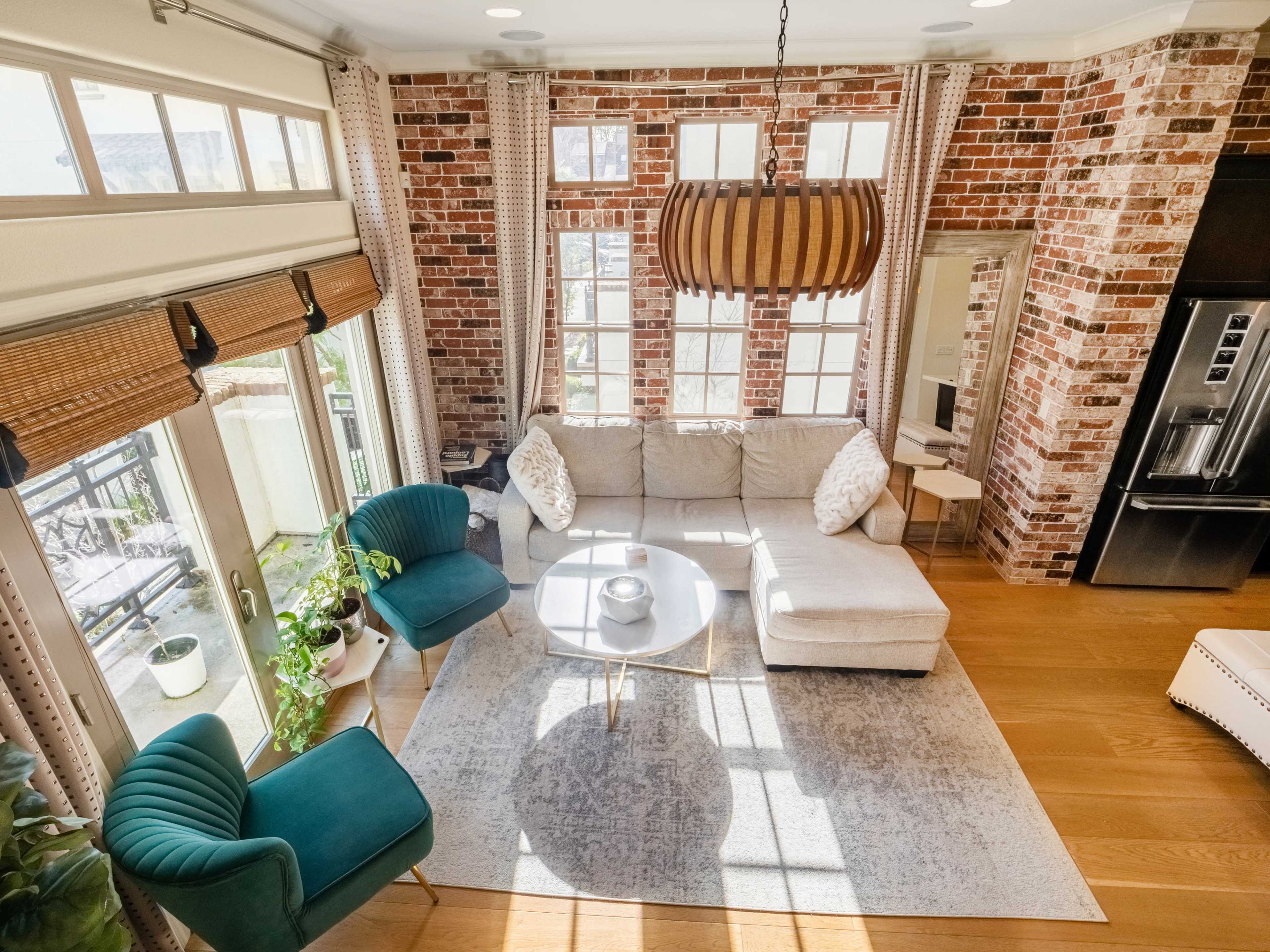 The image shows a cozy living room with a beige couch, two teal chairs, a round coffee table, and large windows that illuminate the space, featuring exposed brick walls and wooden flooring.