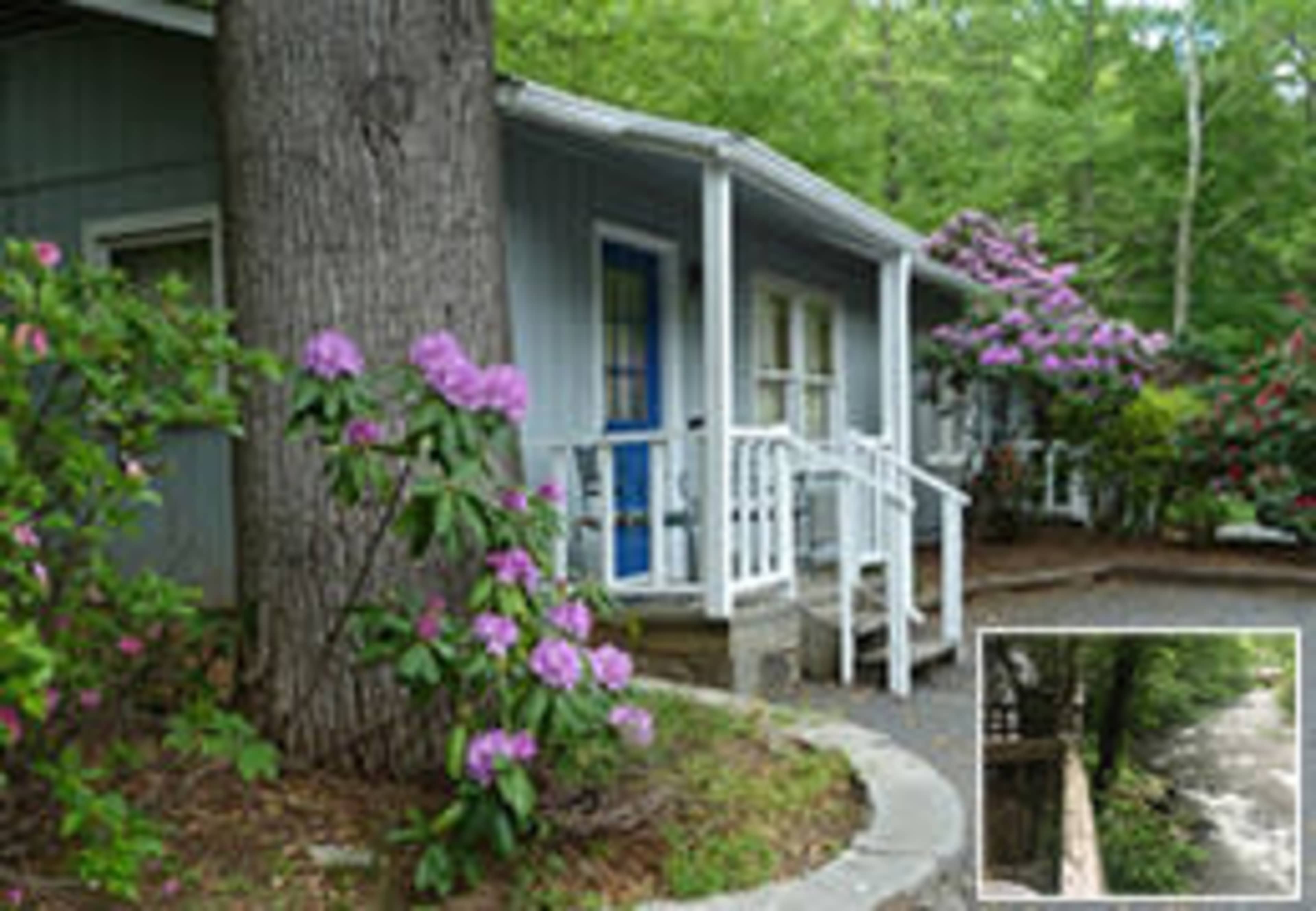 A blue cottage with white railings is surrounded by colorful flowering plants and trees.