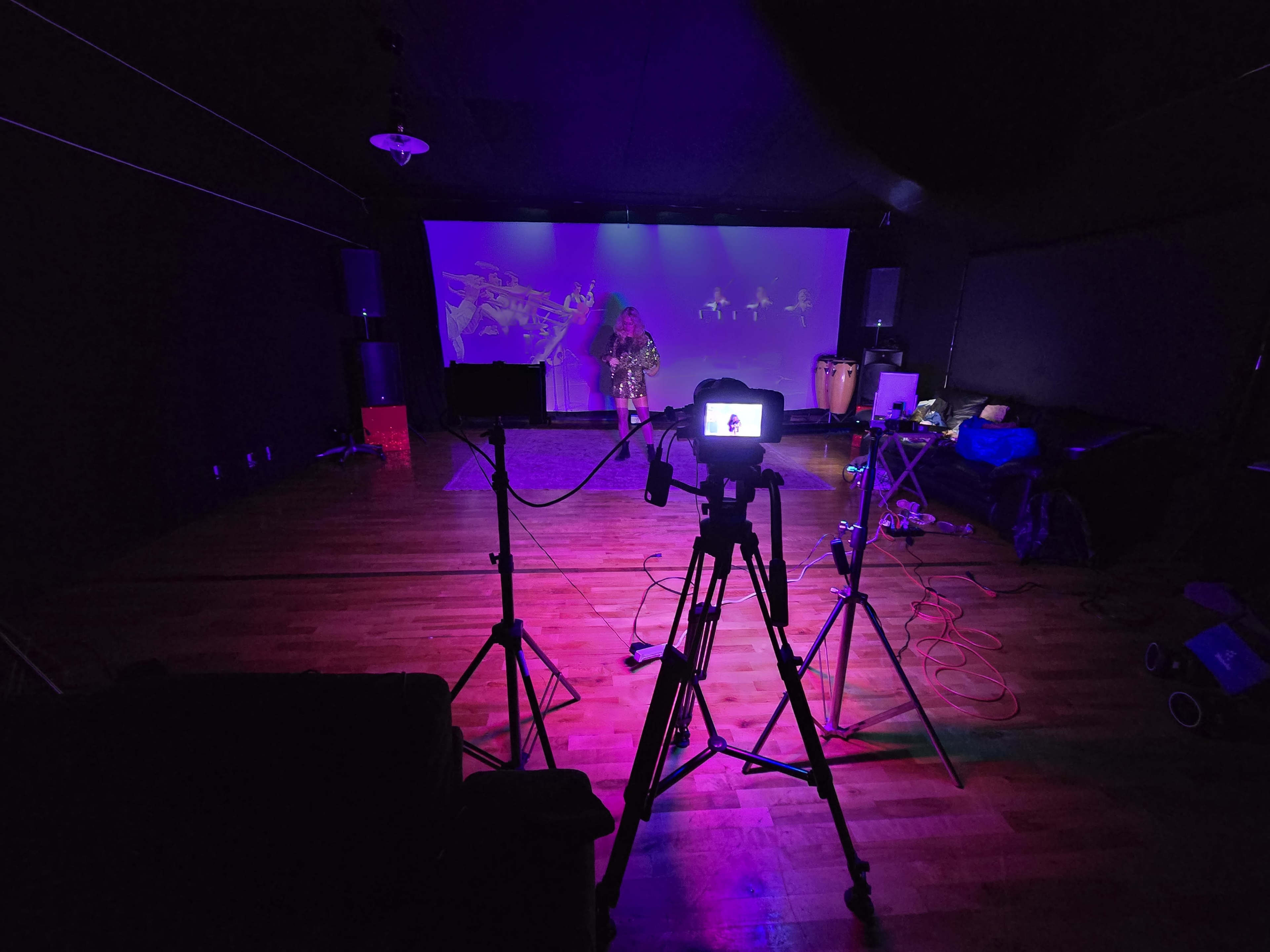 A performer stands in front of a projected screen in a dimly lit room, surrounded by musical equipment and instruments.