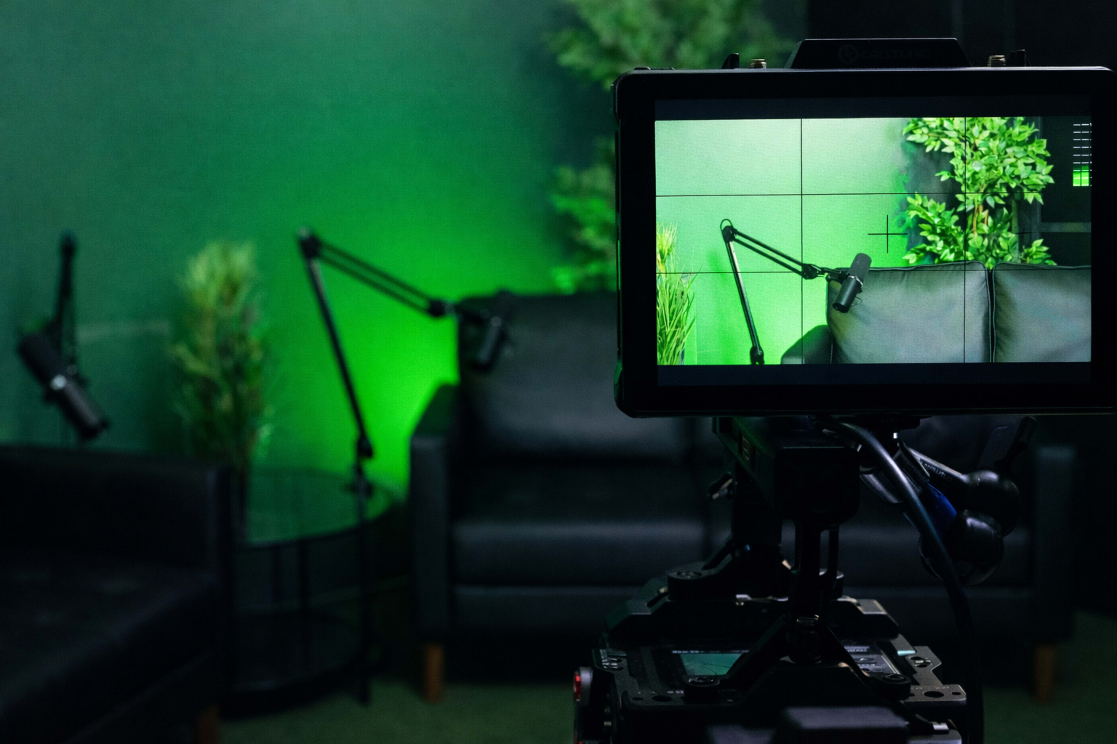 A camera is focused on a seated area with dark sofas and green lighting, surrounded by leafy plants and microphone stands.