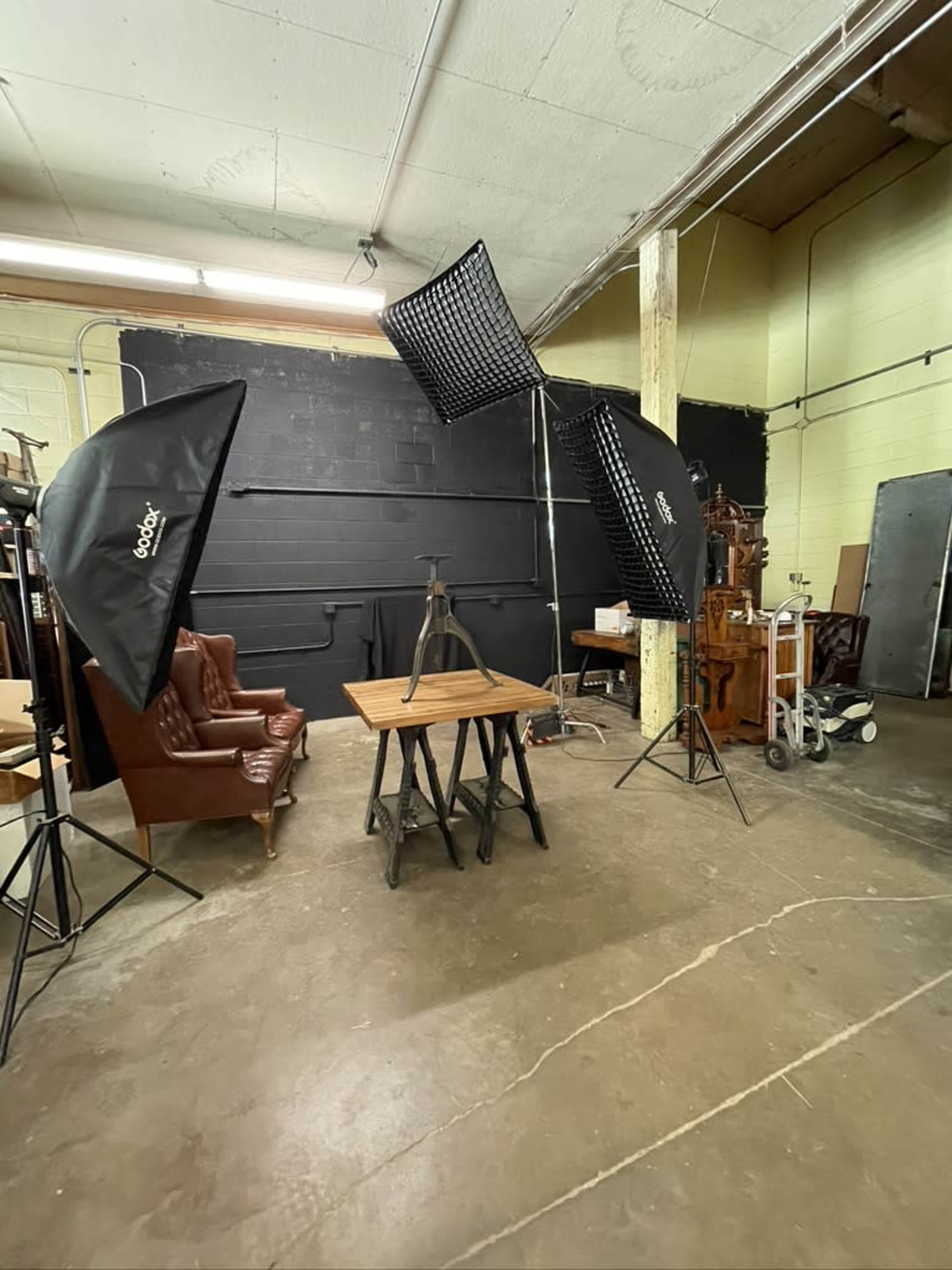 The image shows a photography studio setup with two large softbox lights, a wooden table on a metal stand, and a leather armchair in the background.