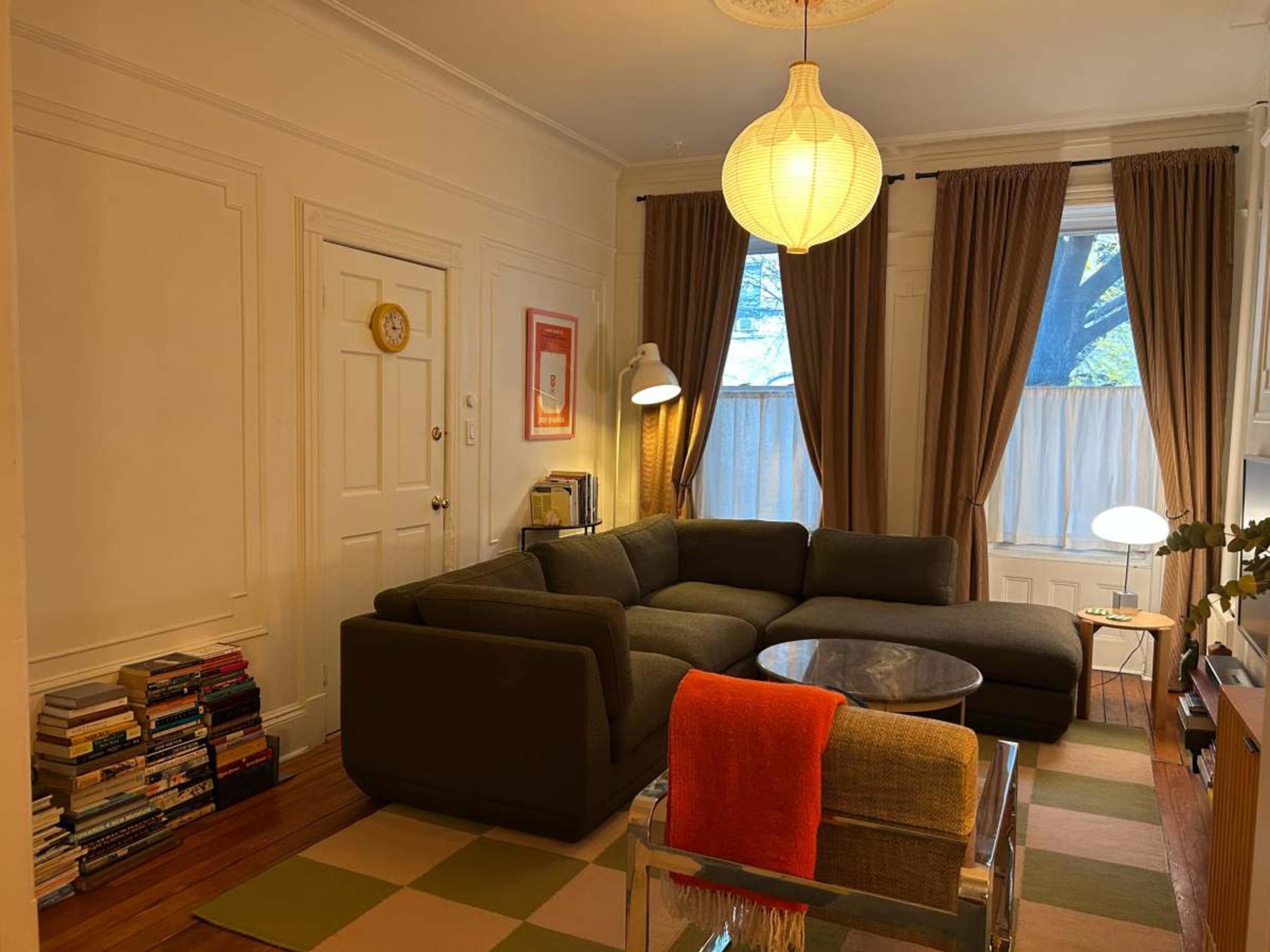 The image features a cozy living room with a sectional sofa, a coffee table, and a bookshelf filled with books, complemented by large windows with curtains.