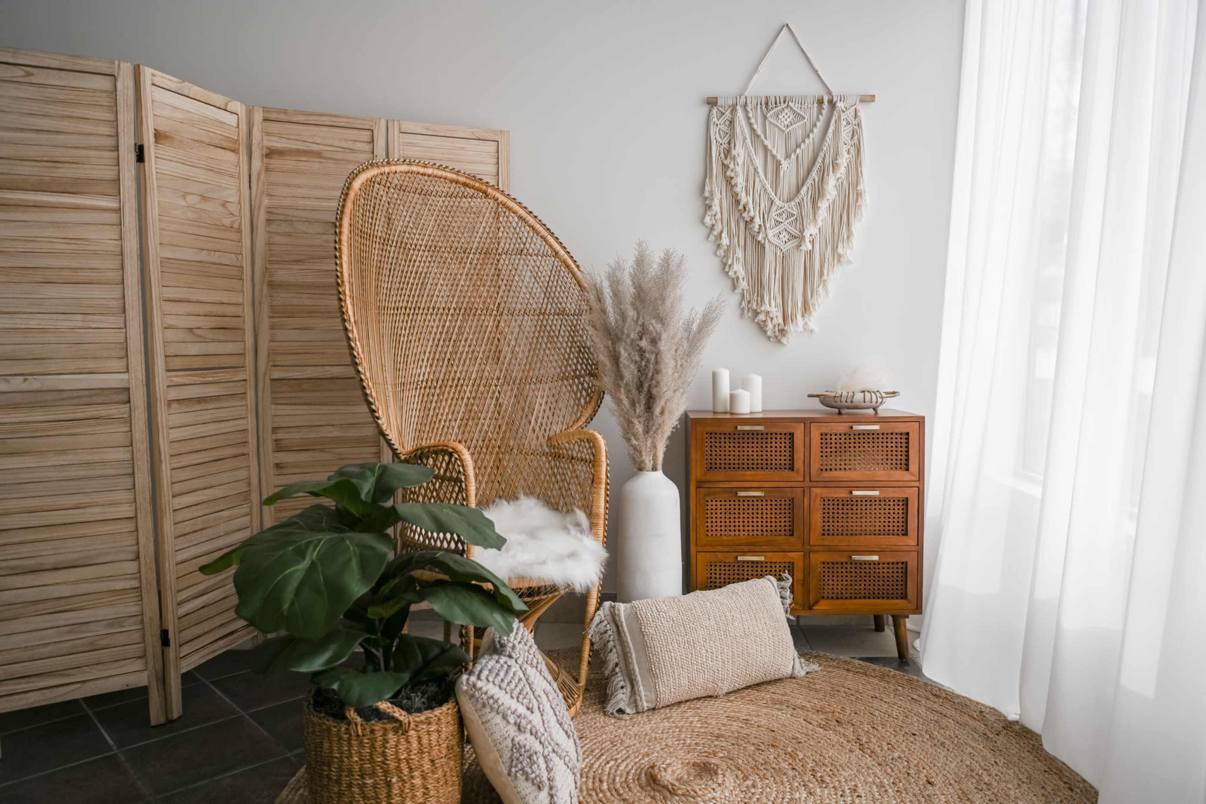 A cozy interior space featuring a large rattan chair, decorative wall hanging, potted plant, patterned throw pillows, and a wooden storage unit with woven details.