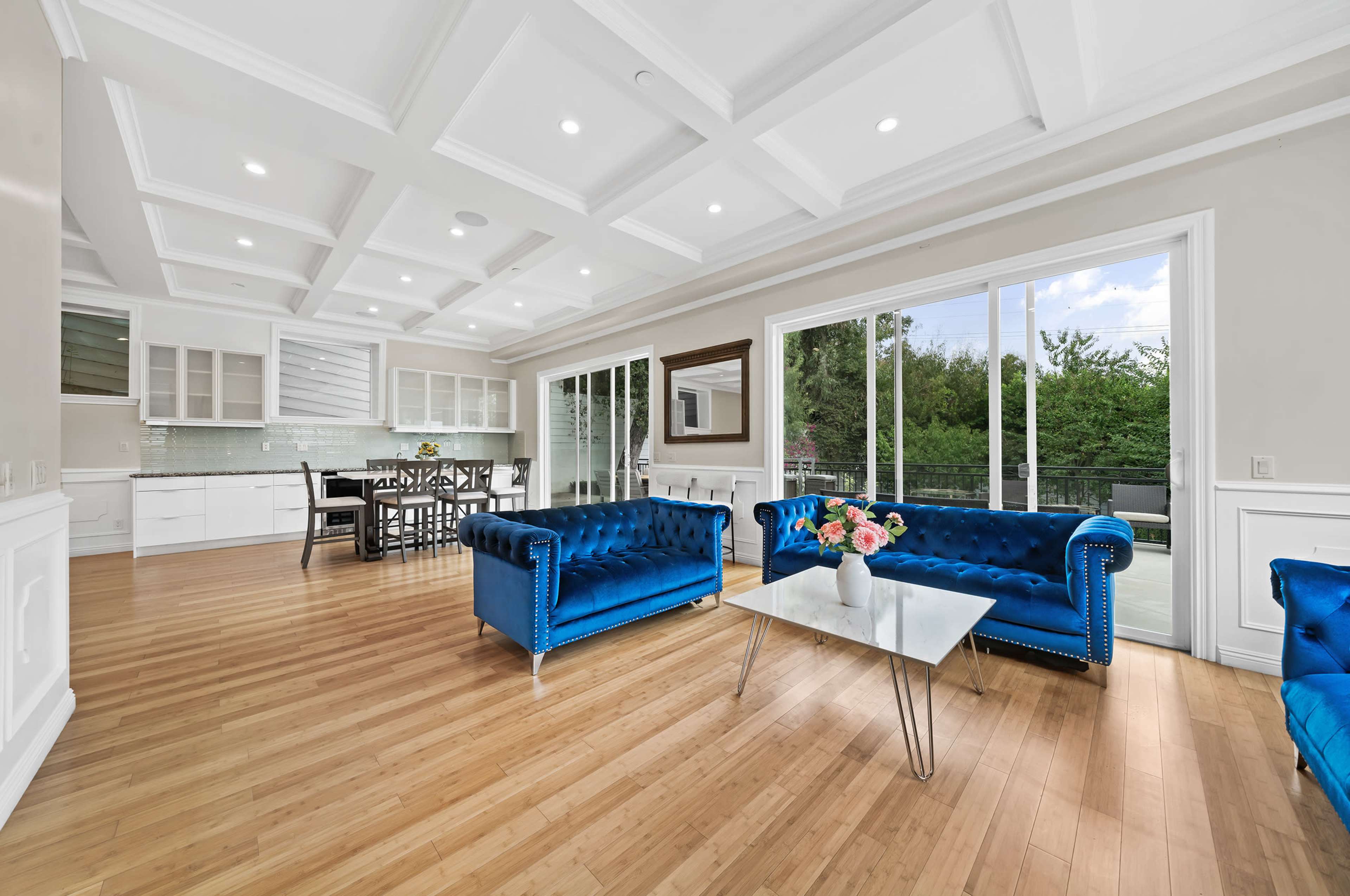 The image shows a spacious living area featuring two blue velvet sofas, a glass coffee table, and a dining space in a house with large windows allowing natural light to fill the room.