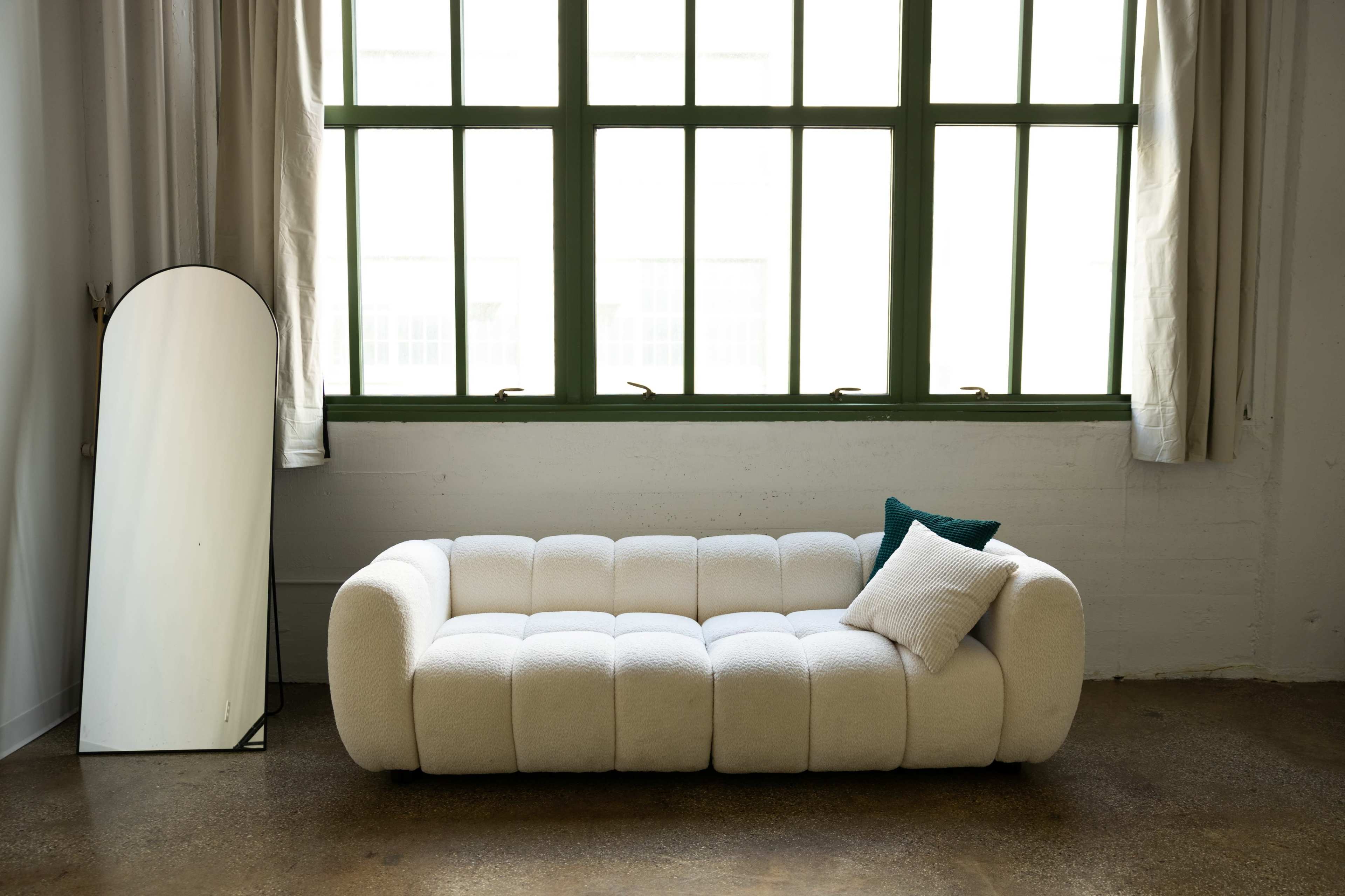 The image shows a white, tufted sofa positioned near large windows with green frames, accompanied by a tall, curved mirror.