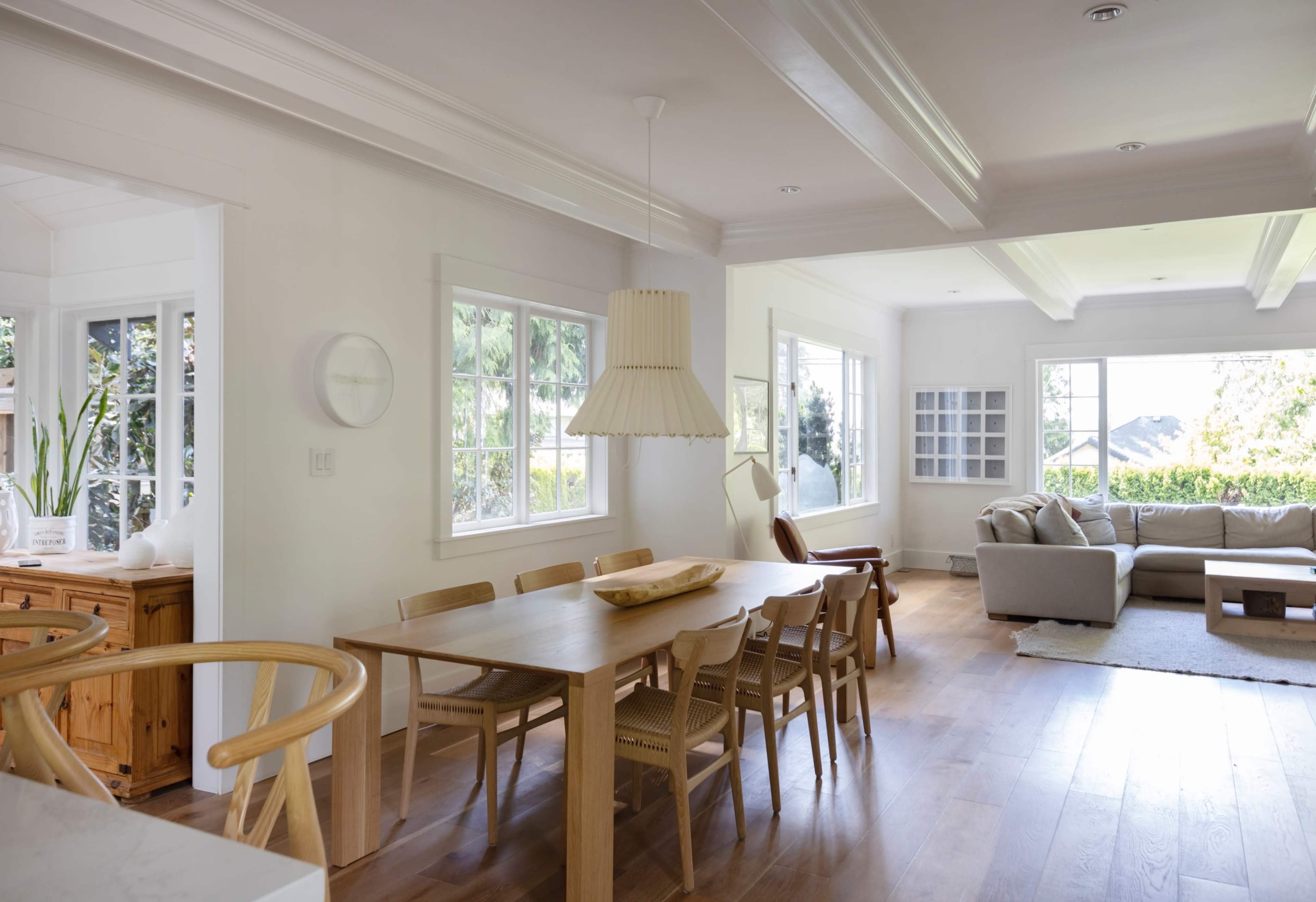 A bright, open-plan living and dining area with a wooden table, chairs, and a sofa, surrounded by large windows.