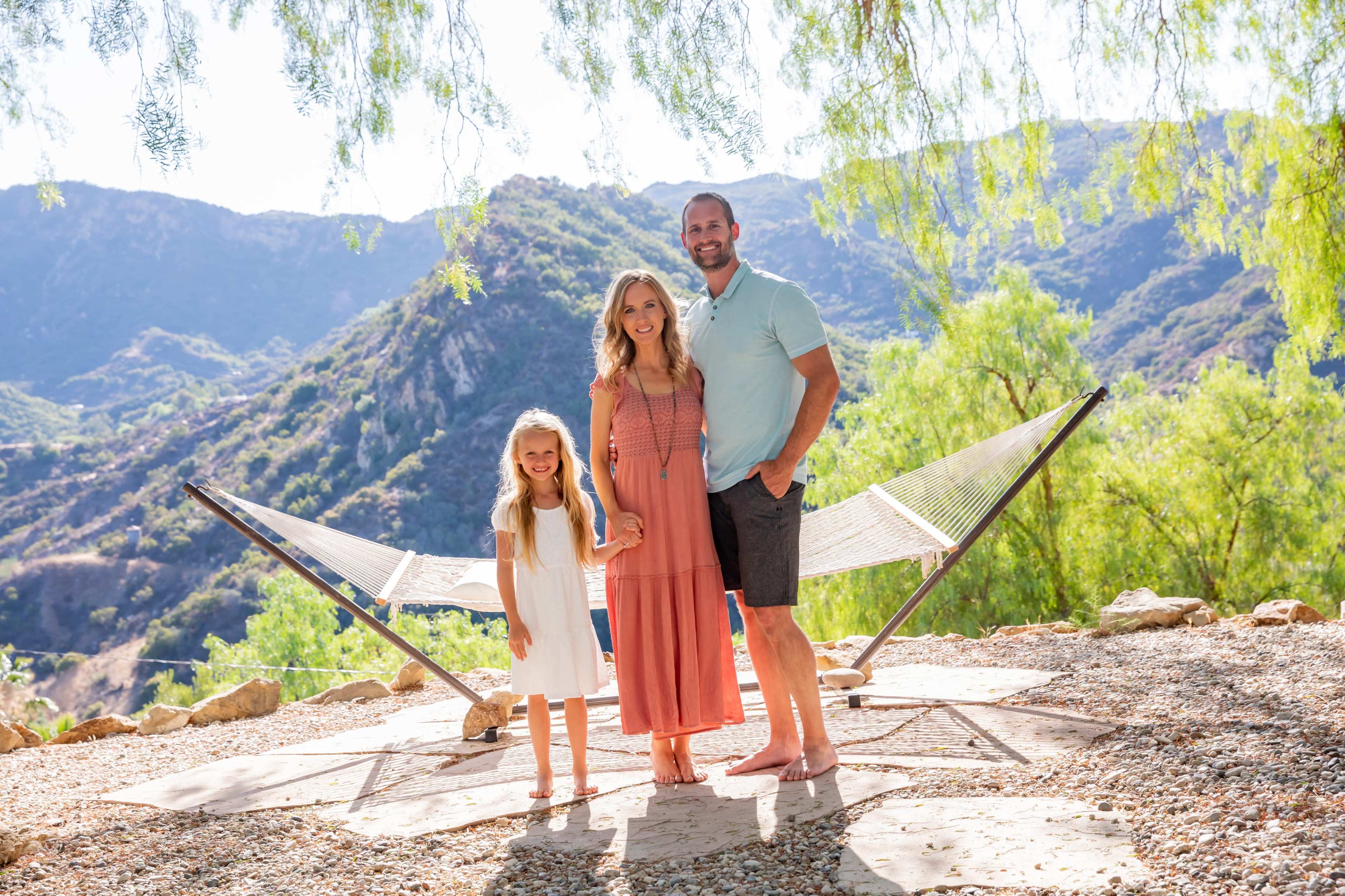 A family of three stands on a gravel area in front of a hammock, with green trees and mountains in the background.