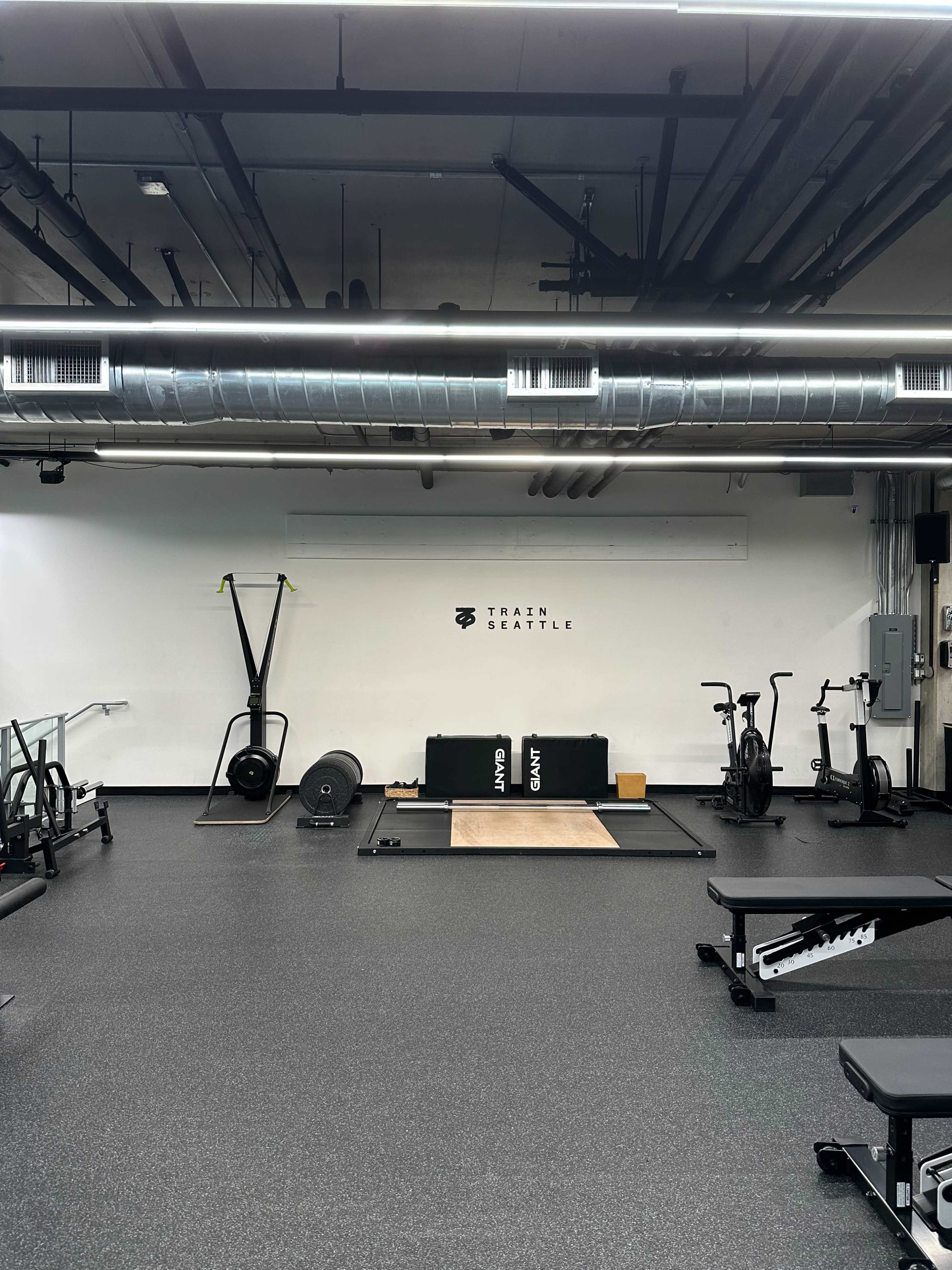 The image shows a modern gym interior featuring various exercise equipment, including a weightlifting platform, a sled, and stationary bikes, set against a minimalist white wall with the text "TRAIN SEATTLE."