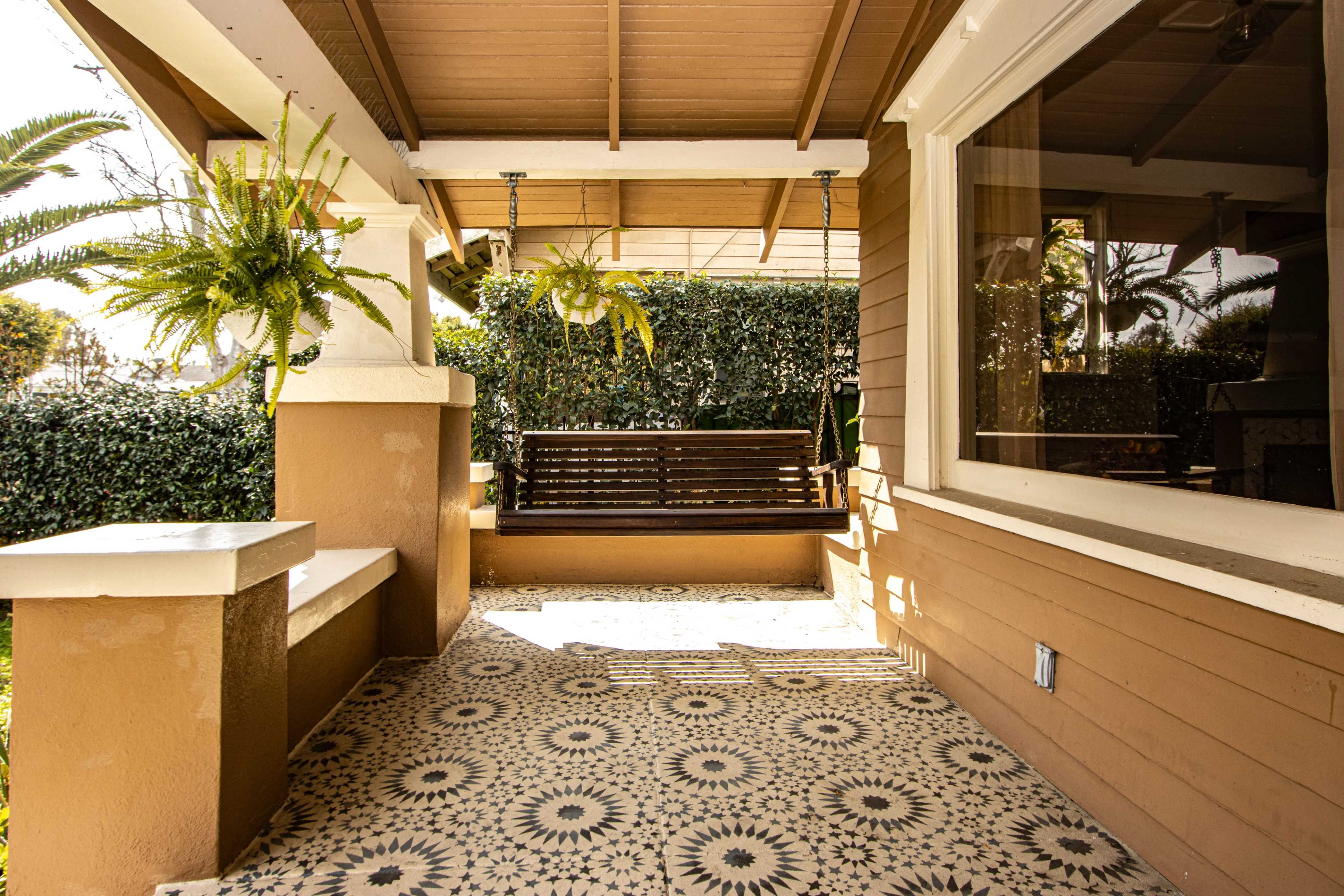 A porch with a patterned floor, a wooden swing, and greenery in the background.