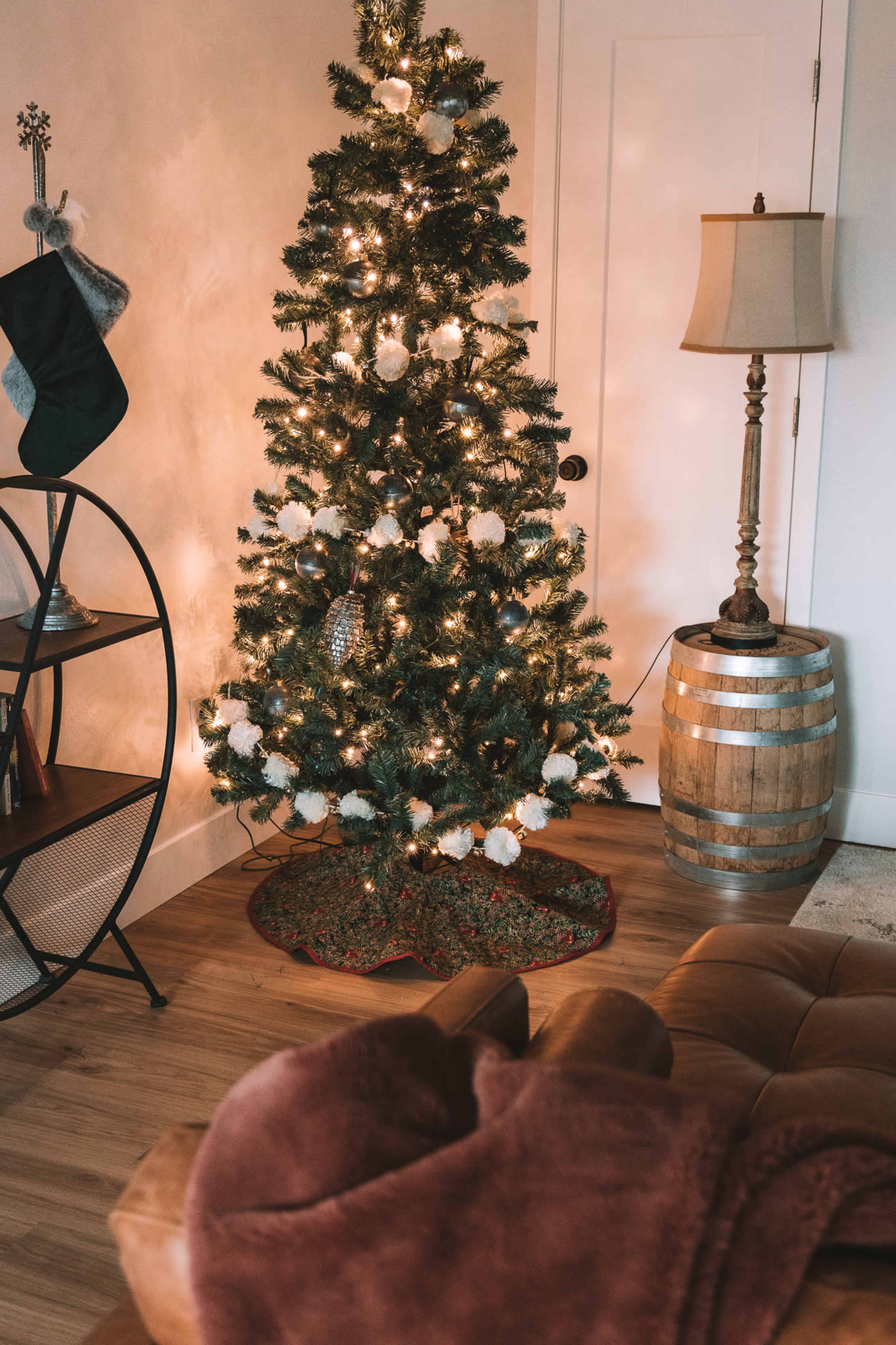 A Christmas tree decorated with lights and ornaments stands in a cozy corner next to a lamp and a wooden barrel.