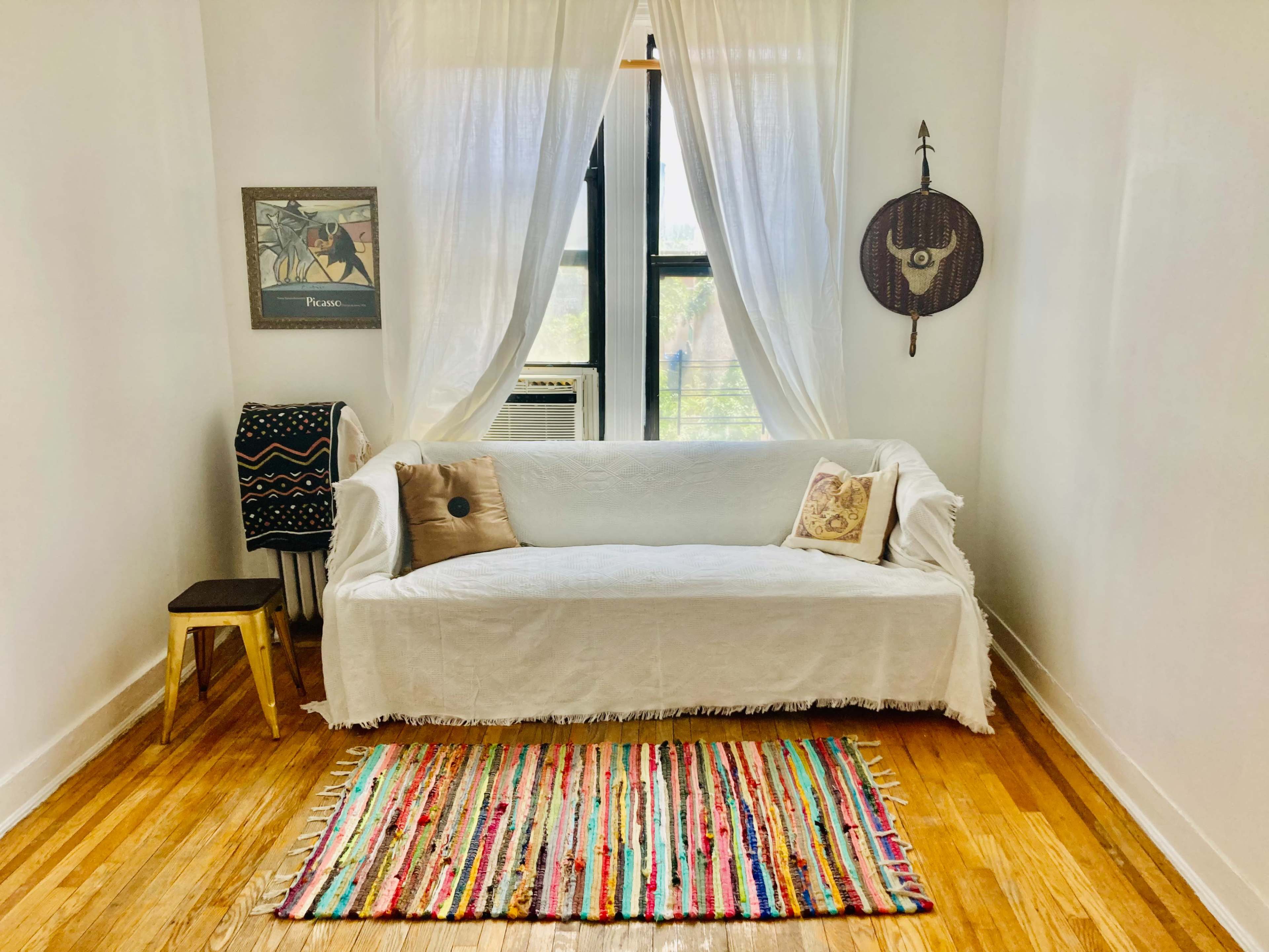 A white couch is draped with a textured covering and sits against a bright wall near a small wooden stool and a colorful woven rug.