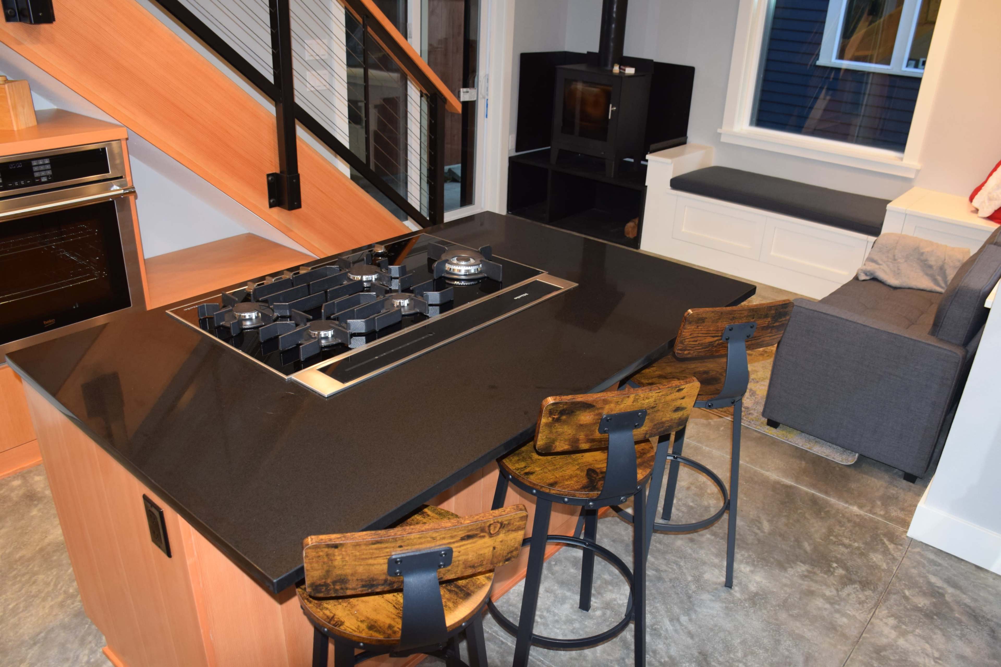 The image shows a modern kitchen with a central island featuring a stovetop, surrounded by three bar stools, and a cozy seating area in the corner.