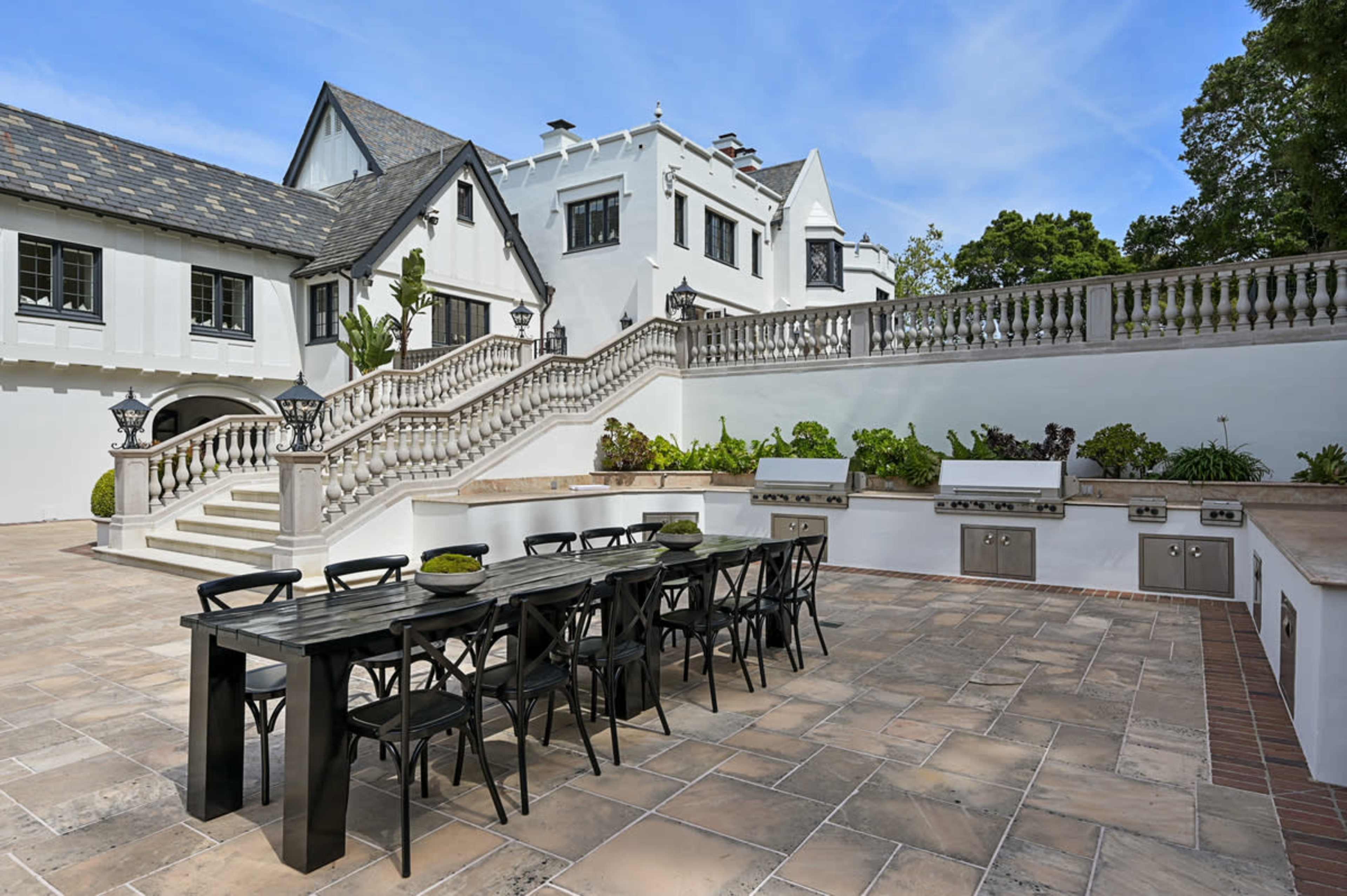 The image shows a large outdoor dining area with a long black table and chairs, situated beside an elegant stone staircase leading to a white mansion with a slate roof.