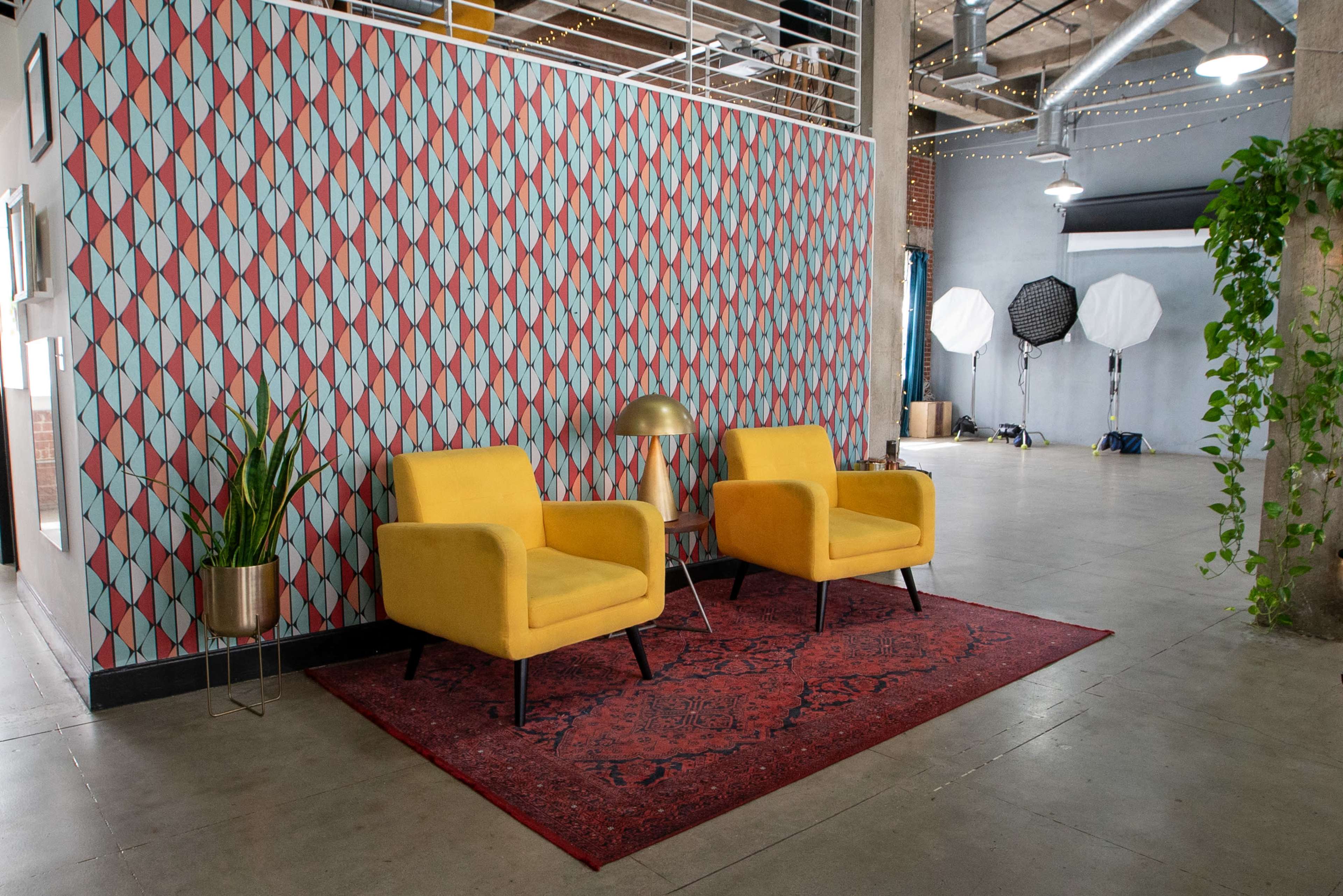 Two yellow chairs placed on a red rug in front of a patterned wall, with lighting equipment and plants in the background.
