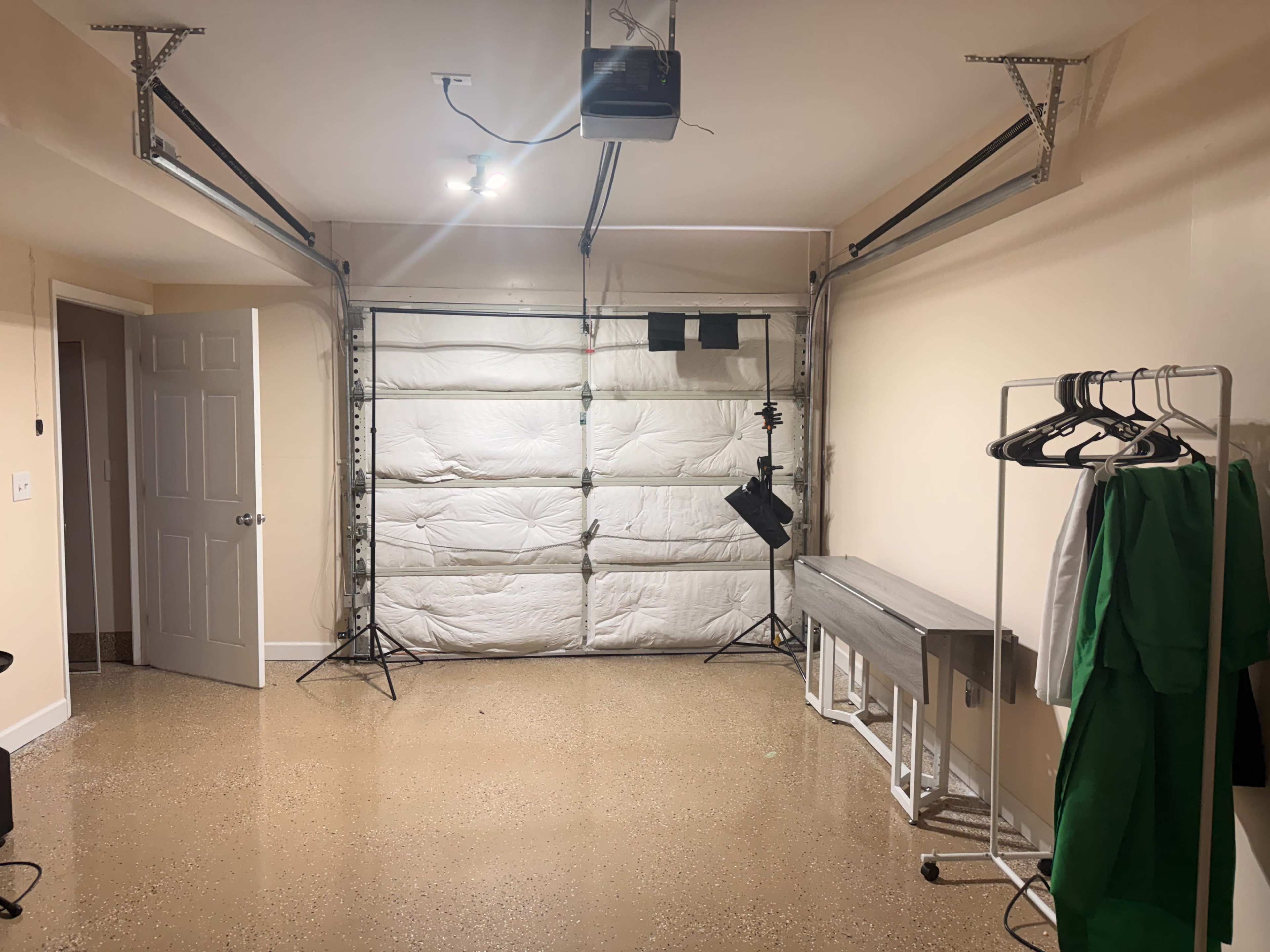 The image shows an empty garage with a closed door, overhead lights, and a few pieces of equipment including a clothing rack and a workbench.