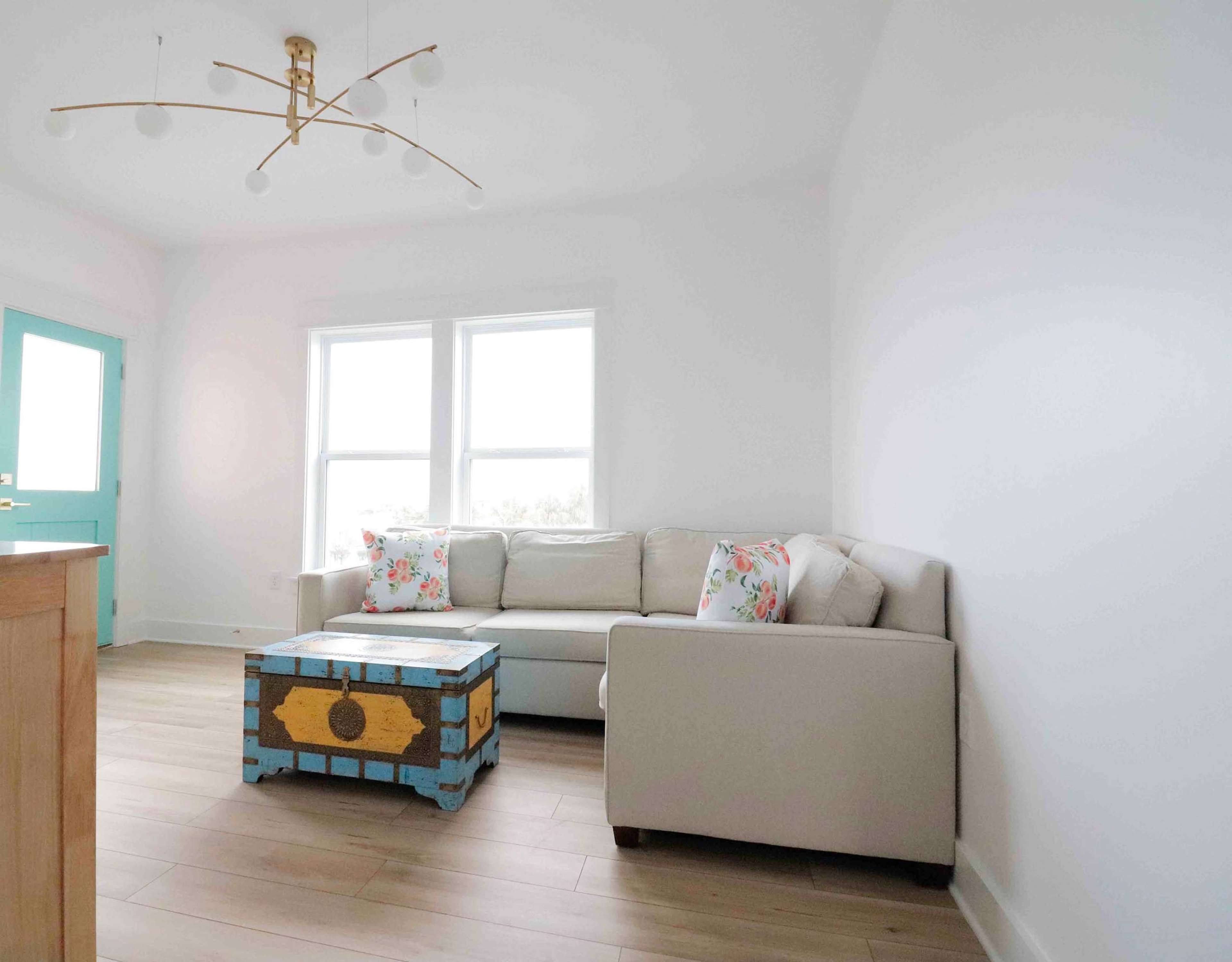 A light-filled living room features a beige sofa with decorative pillows and a colorful coffee table, accompanied by a door painted in teal.