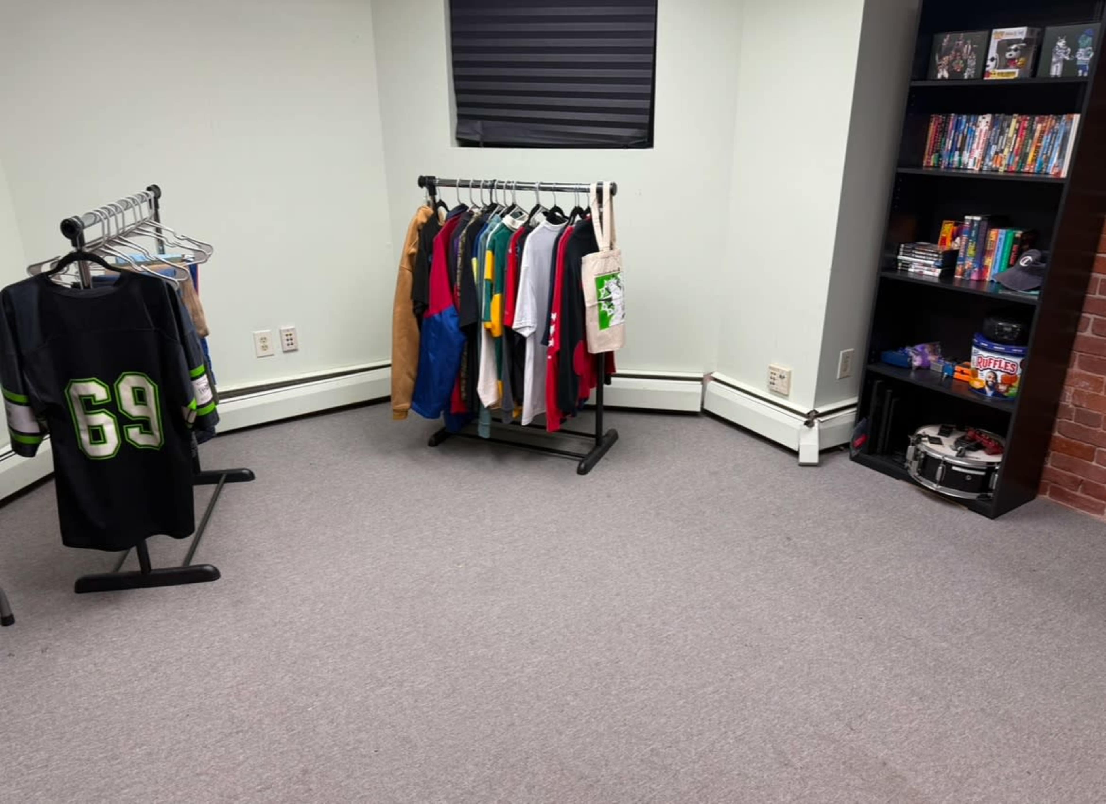 The image shows a room with a clothing rack filled with various shirts and a bookshelf containing DVDs and other items.