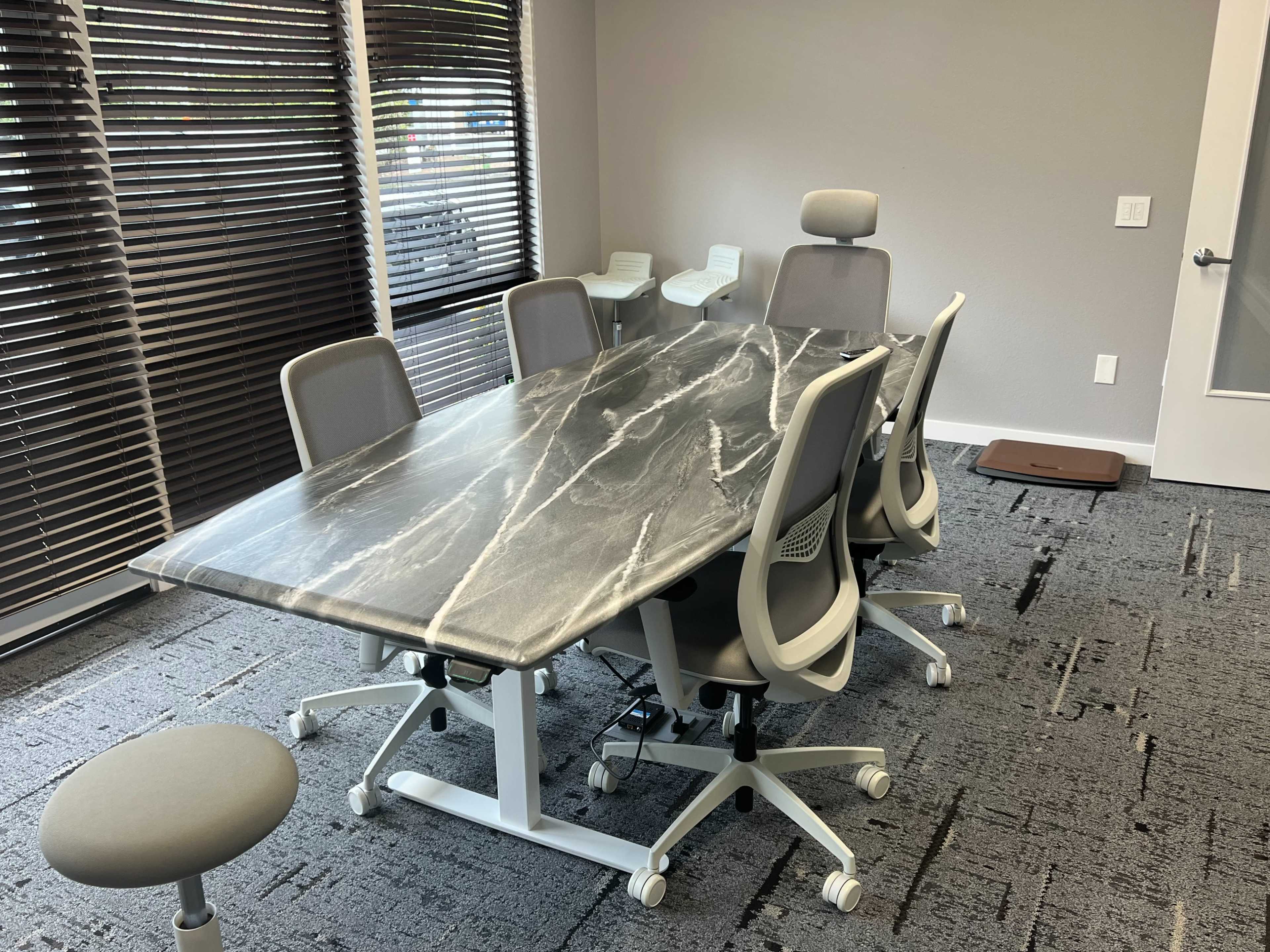 A conference room with a marble-patterned table surrounded by six ergonomic chairs, along with a stool and a window with blinds.
