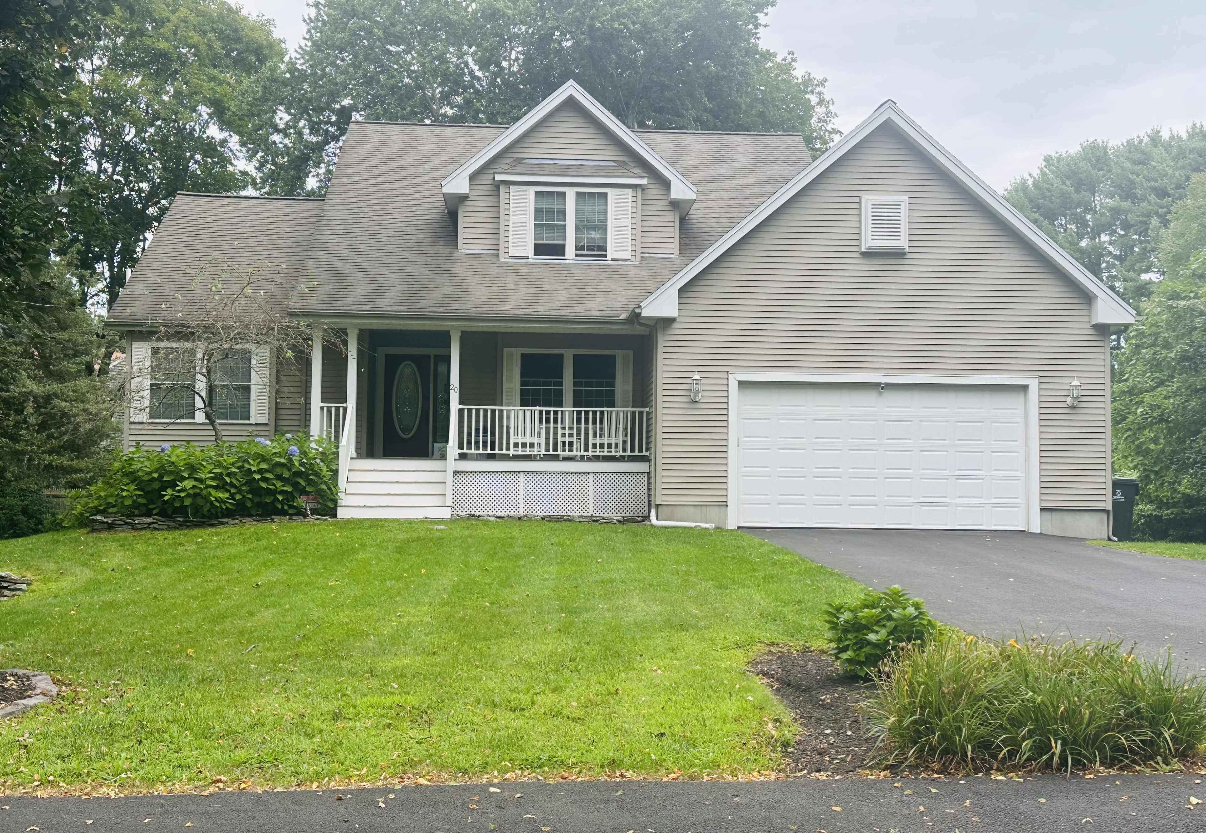 A two-story house with a peaked roof, front porch, and attached garage is situated on a green lawn surrounded by trees.