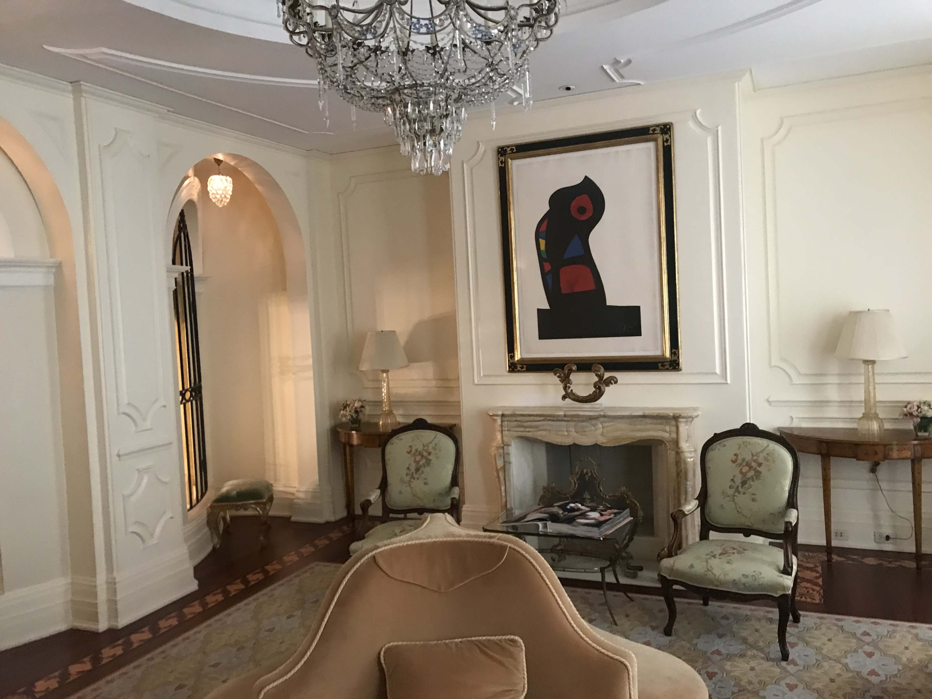 The image shows an elegantly designed room featuring a chandelier, a framed artwork, and two floral-patterned armchairs arranged near a fireplace.