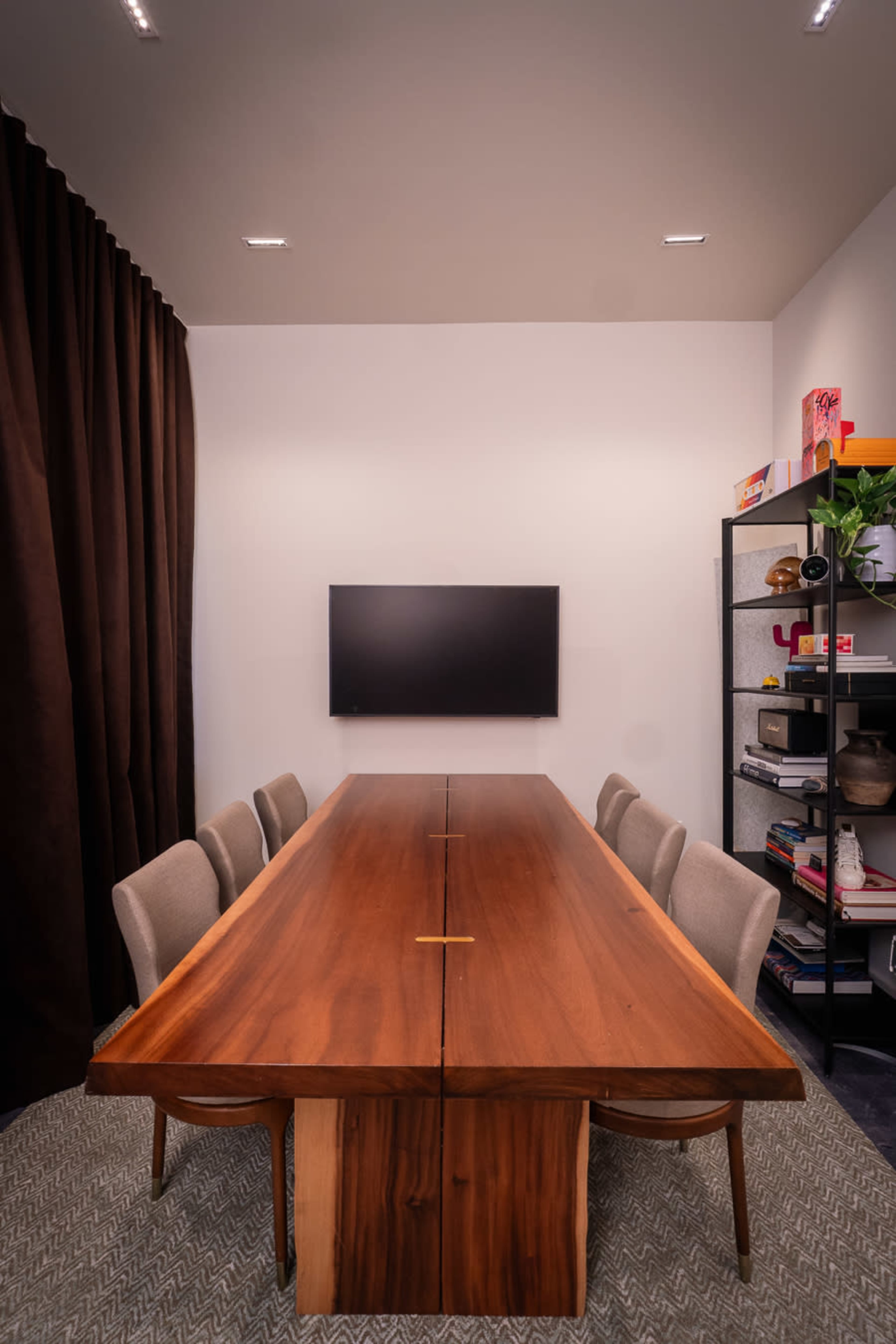 The image shows a modern conference room featuring a long wooden table, surrounded by beige chairs, with a television mounted on the wall and a shelving unit filled with decorative items.