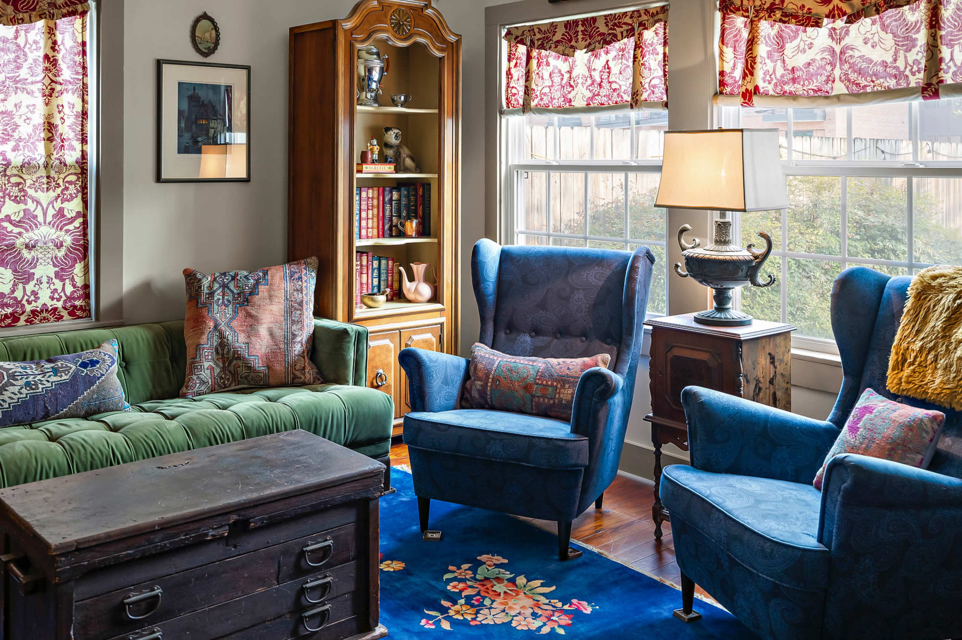 The living room features a green sofa, two blue armchairs, a wooden cabinet, and large windows with floral curtains.