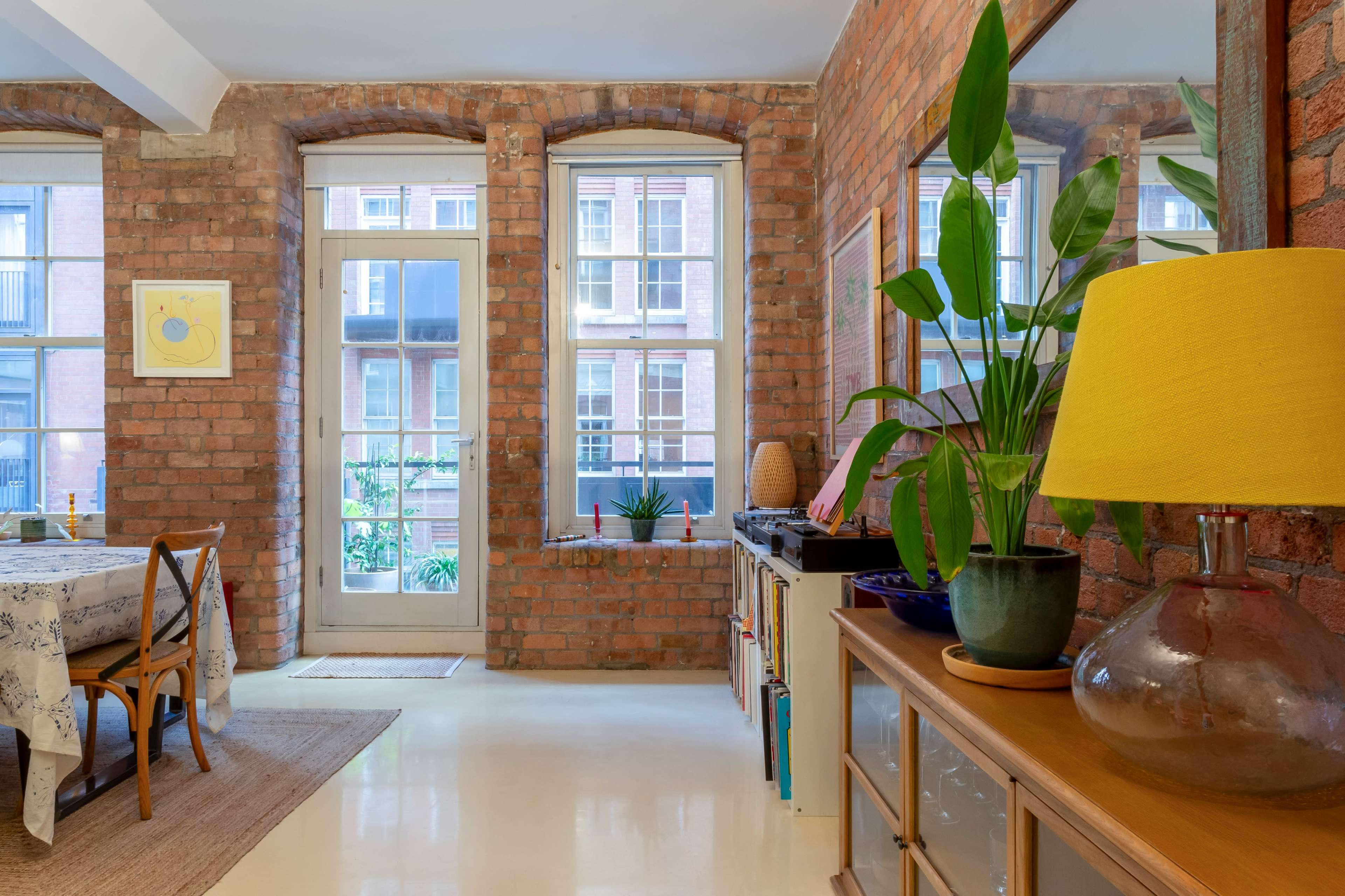The scene shows a bright, open interior featuring exposed brick walls, large windows, a dining table, a vibrant lamp, and greenery.