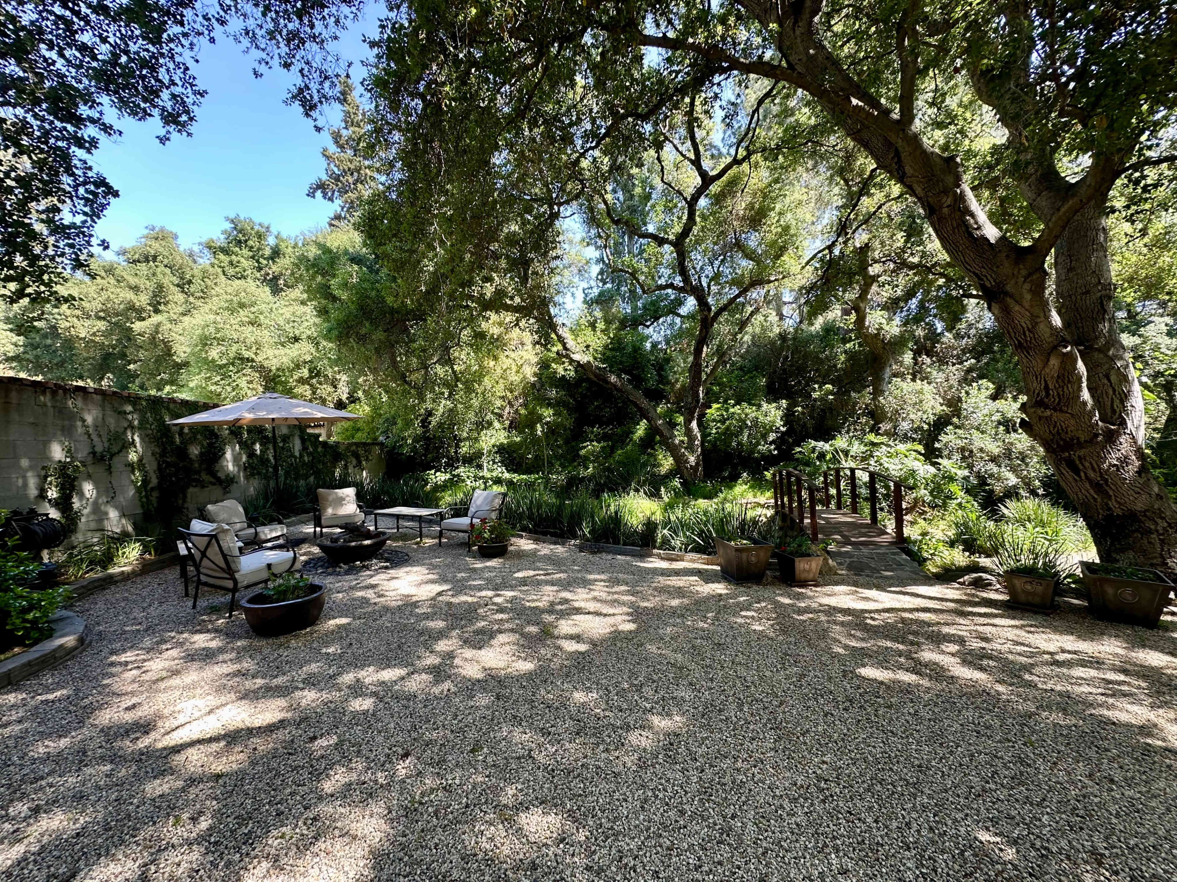 The image shows a gravel courtyard with seating, surrounded by trees and greenery, and a wooden bridge in the background.