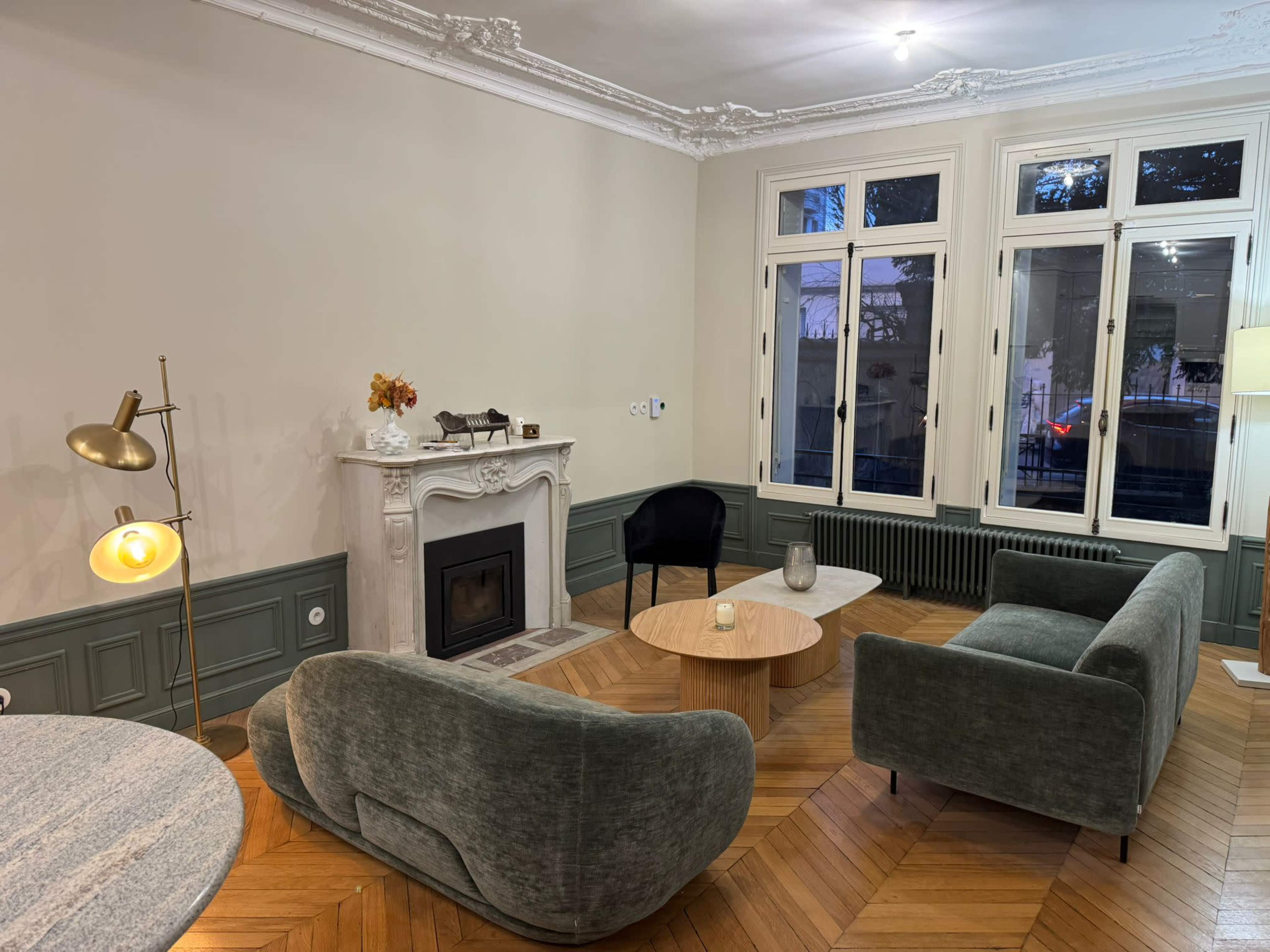 The image shows a well-furnished living room featuring two plush sofas, a round coffee table, and a fireplace set against a backdrop of large windows.