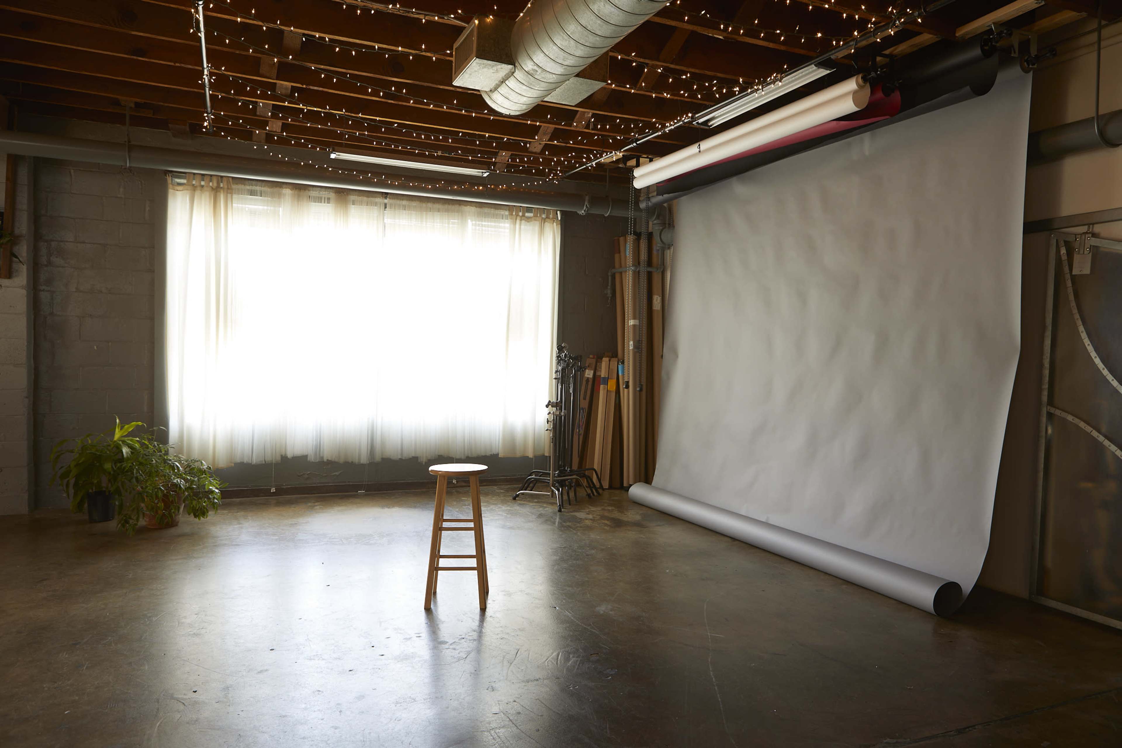 A photography studio features a wooden stool in front of a large gray backdrop, illuminated by natural light from a window.