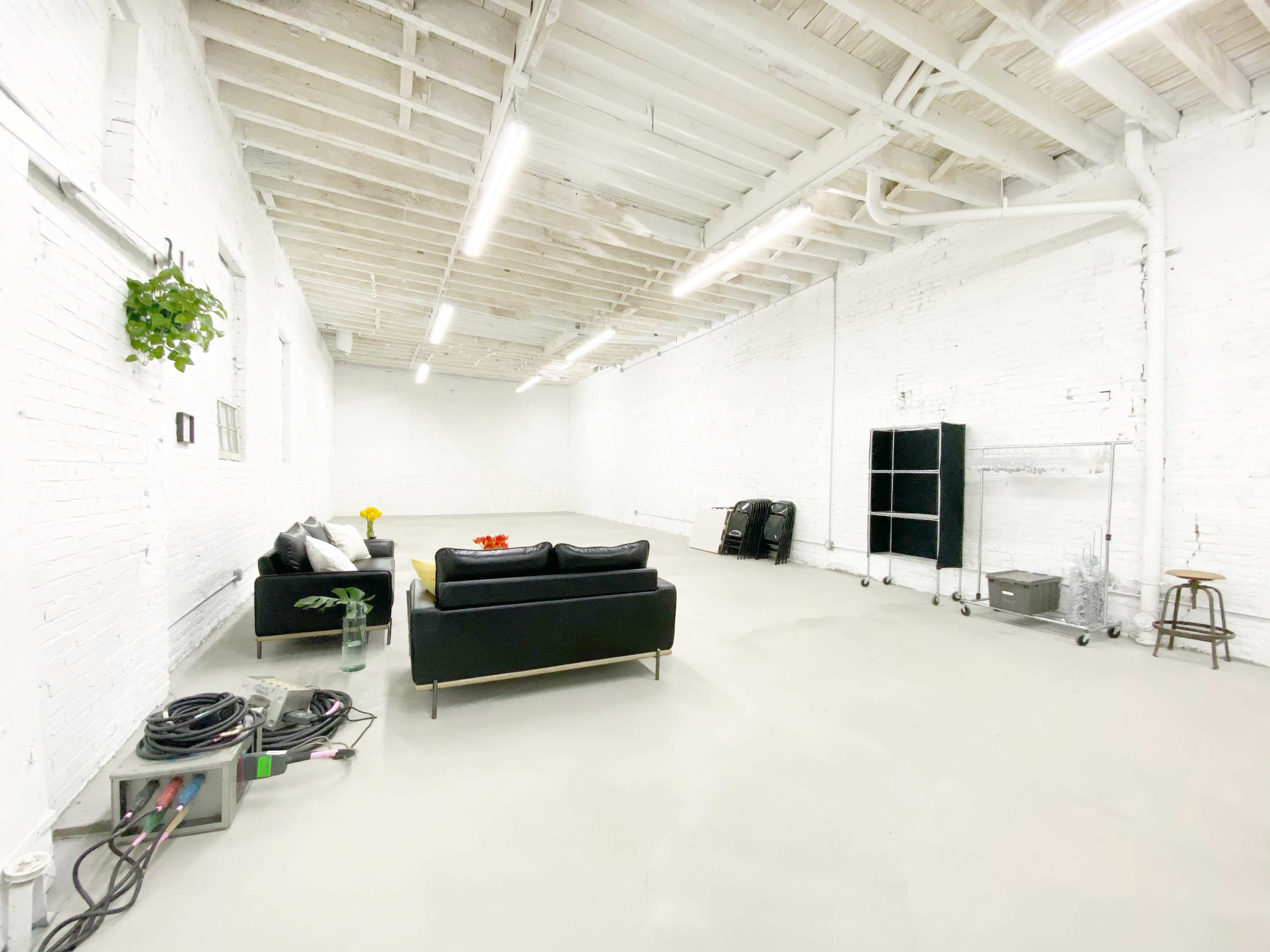 A spacious, minimally furnished white room with a black sofa, a small plant, and various items on the floor, including cables and furniture pieces.