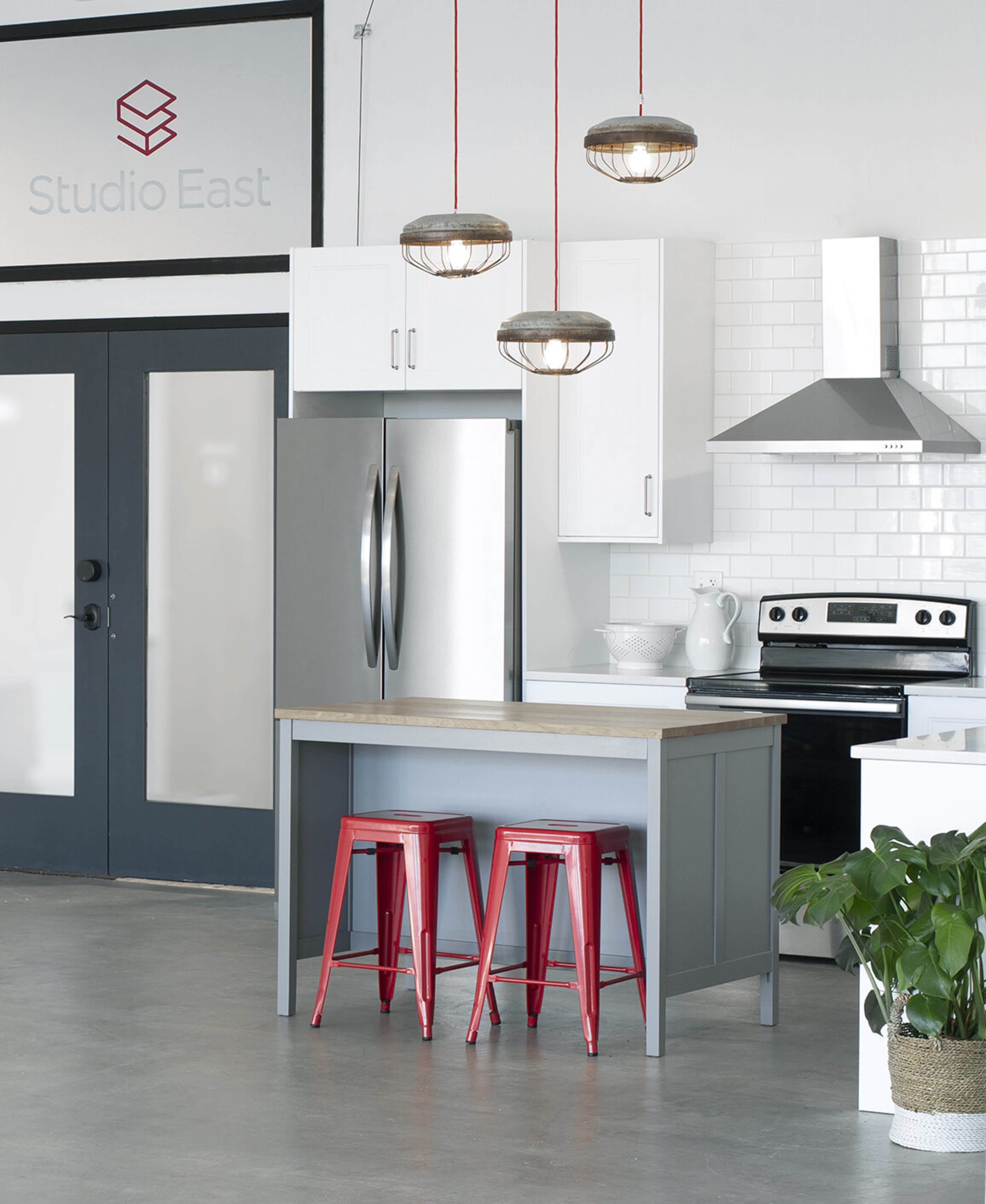 The image shows a modern kitchen with a wooden island, two red metal stools, stainless steel appliances, and pendant lighting.
