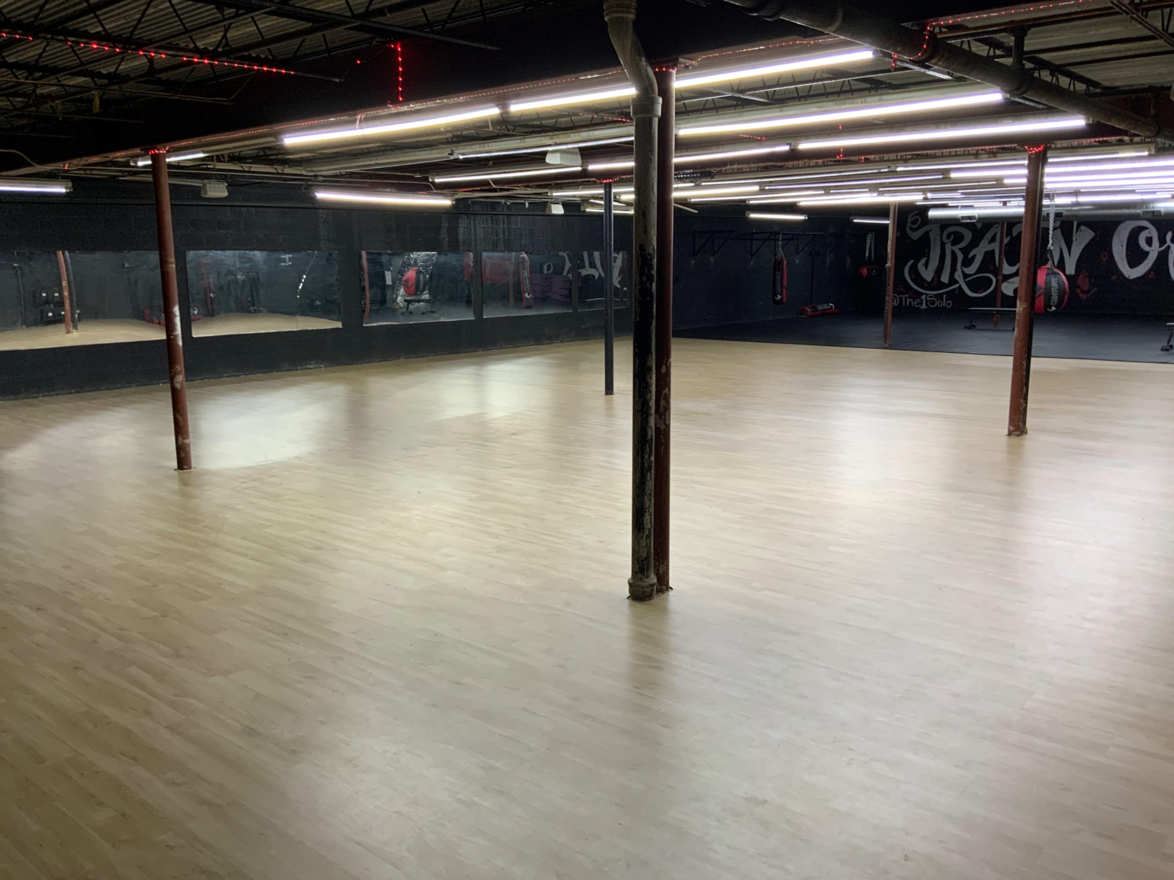 The image shows a large, empty fitness studio with wooden flooring and mirrored walls, illuminated by overhead lights.