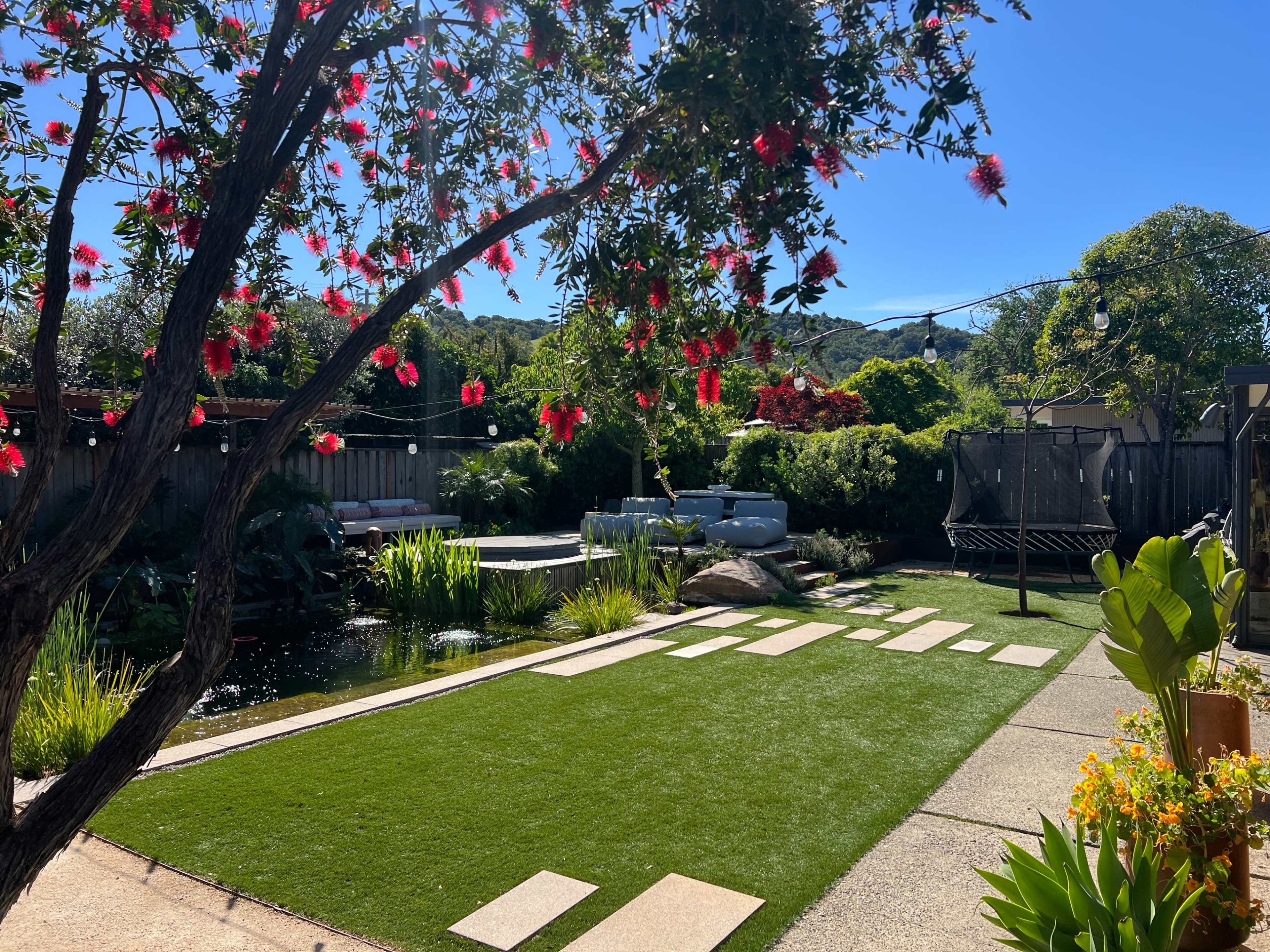 A well-maintained garden features green grass, decorative stones, a water feature, and flowering trees under a clear blue sky.