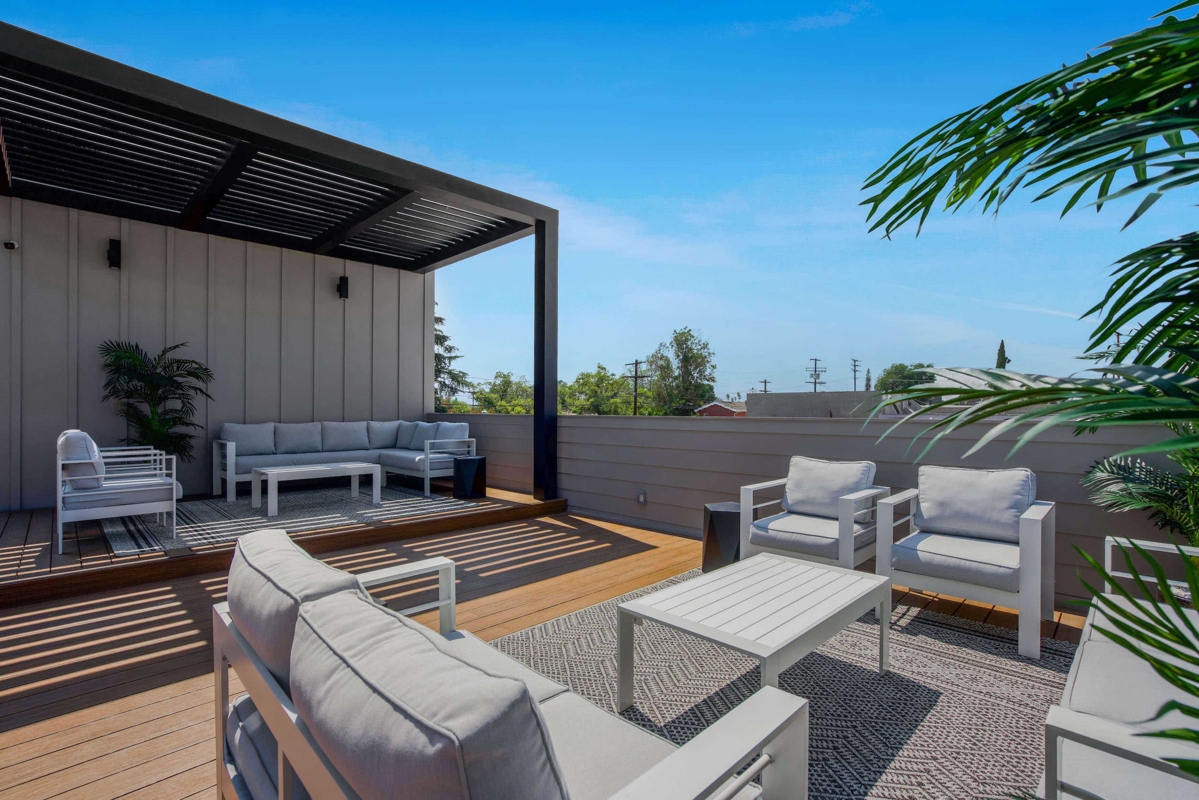 A rooftop patio features a seating area with gray furniture and a patterned rug, surrounded by greenery and a clear blue sky.