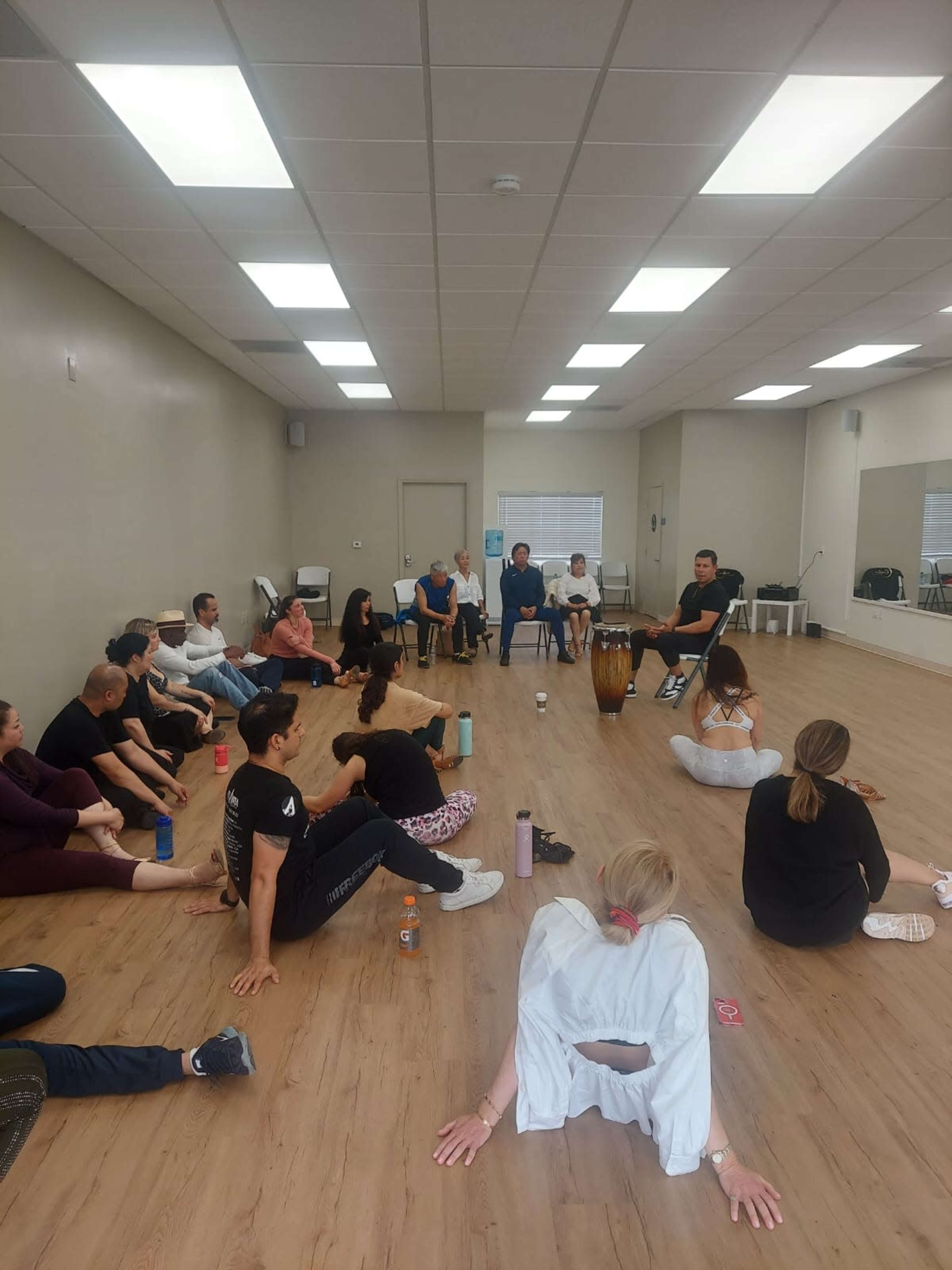 A group of people is seated in a dance studio, with some stretching on the floor while others listen to a musician playing a drum.