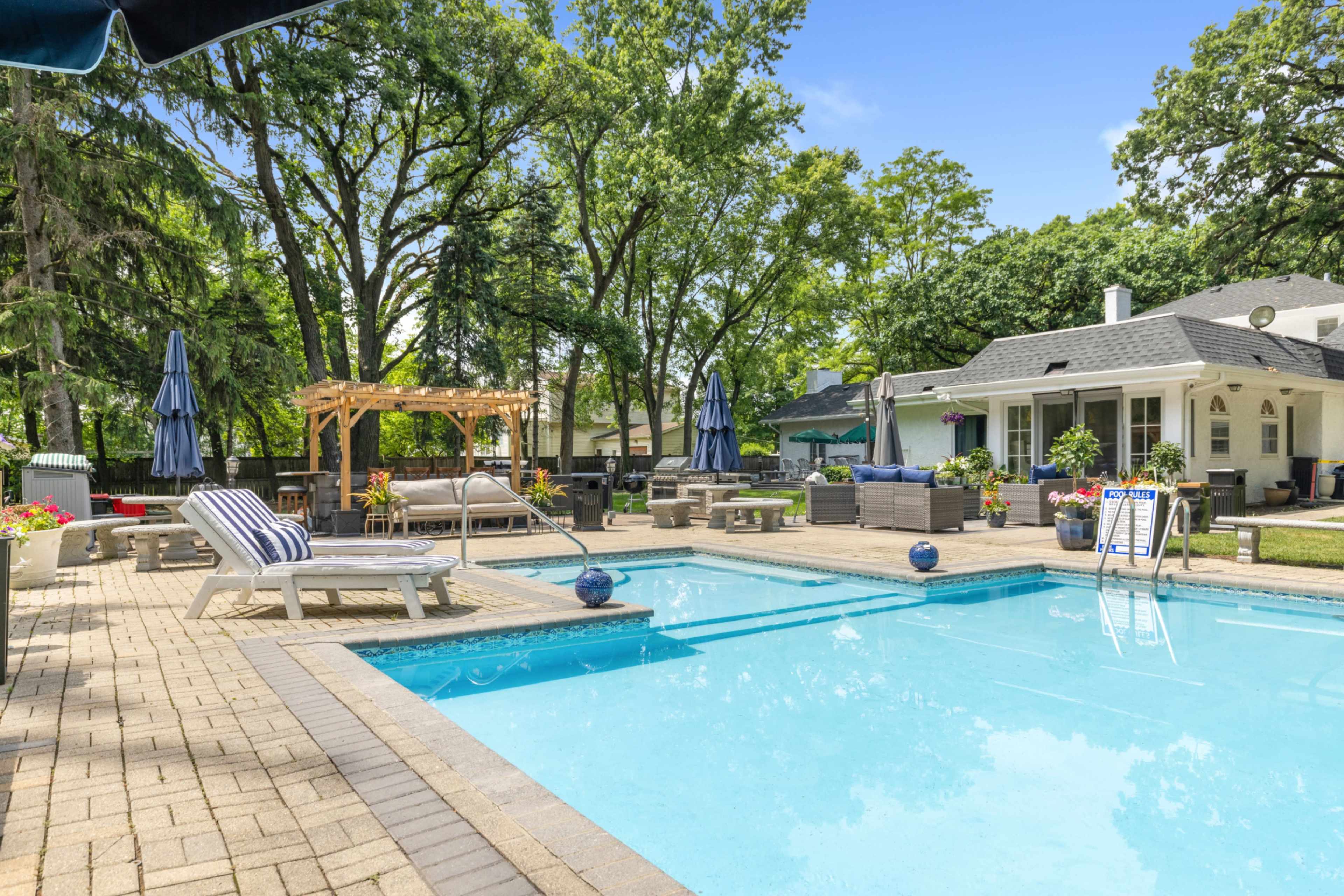The image shows a well-maintained backyard featuring a swimming pool, lounging chairs, a gazebo, and a house surrounded by trees.
