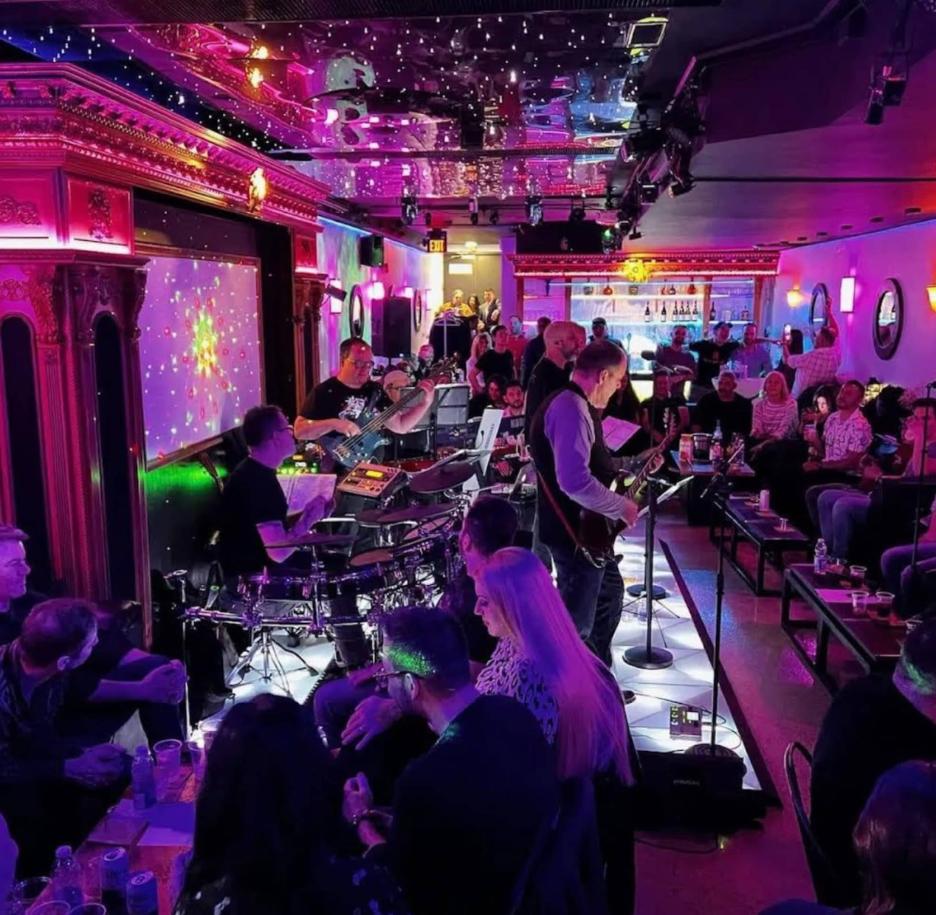 A live band performs on a stage in a brightly lit bar, while an audience sits at tables enjoying drinks.